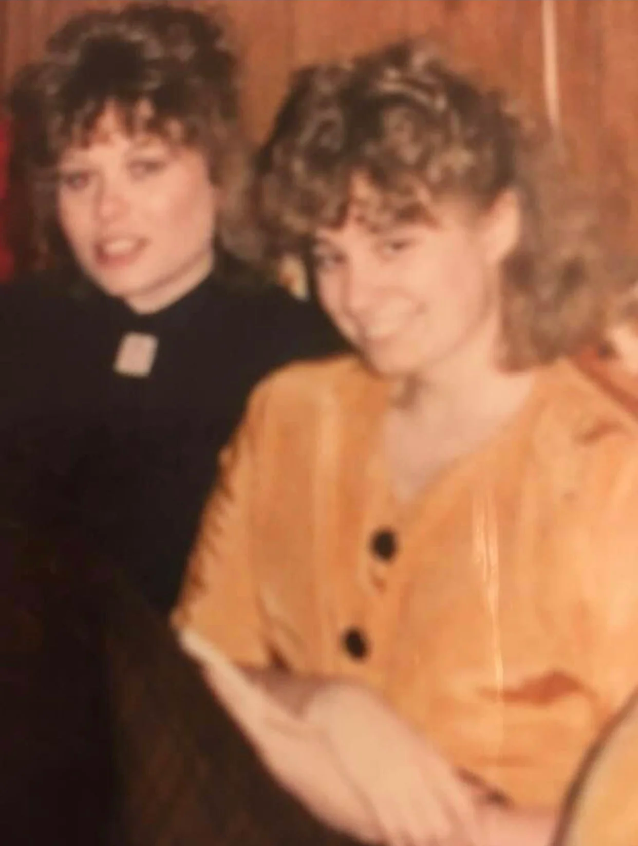 A woman and a teenage girl with curly hair smiling, sitting together in a cozy room. The woman is wearing an orange outfit with buttons, and the girl is wearing a black top with a small white collar.