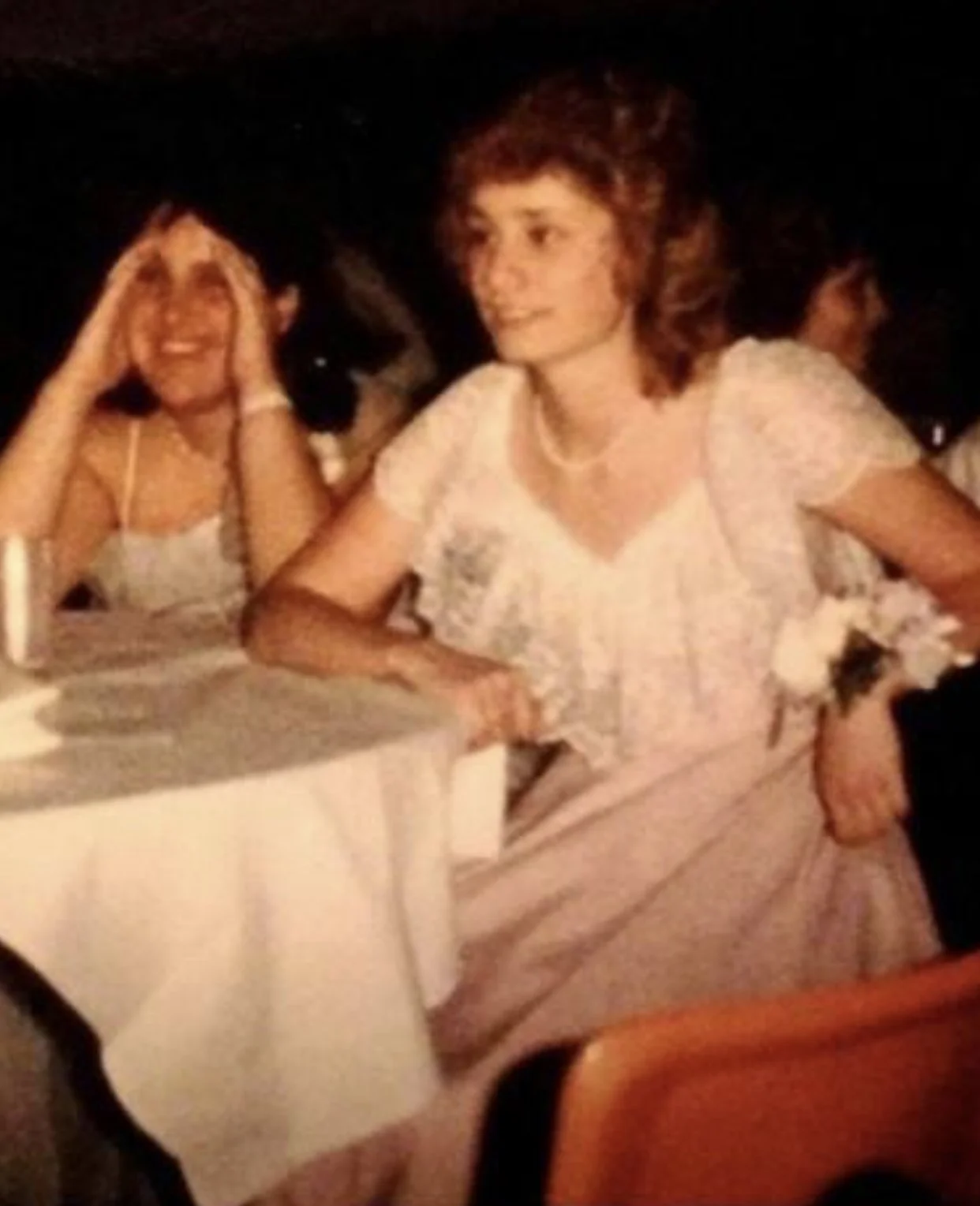A woman with short, curly red hair sitting at a table in a dark room, wearing a white blouse. Another woman with black hair is sitting behind her, smiling, and holding her head with both hands.
