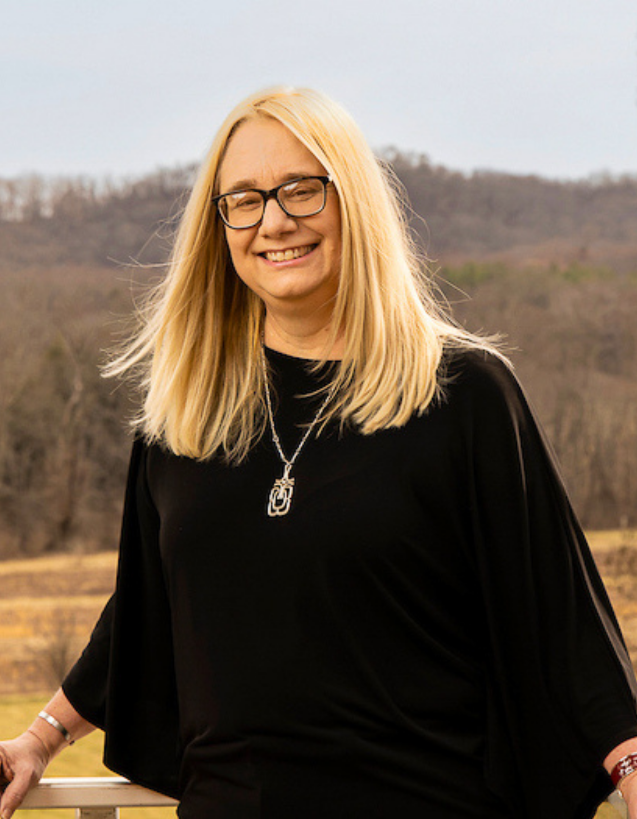 Dreama wearing glasses and a black top, smiling outdoors with a landscape of trees and hills in the background.