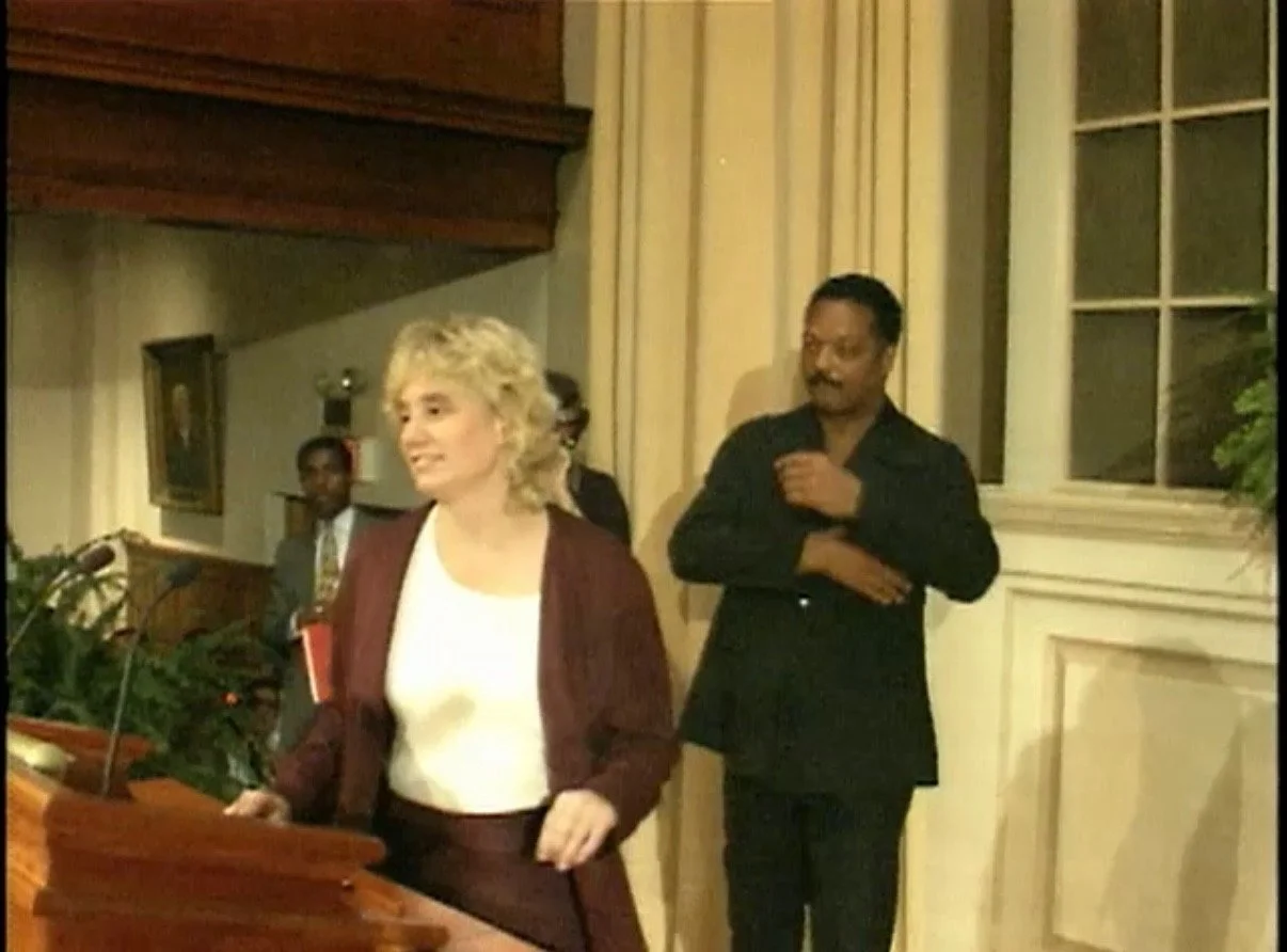 A woman with blonde curly hair in a burgundy suit standing at a podium with a microphone, smiling, with a man in a black suit and tie standing behind her, in a room with cream-colored walls, a window, and some plants.