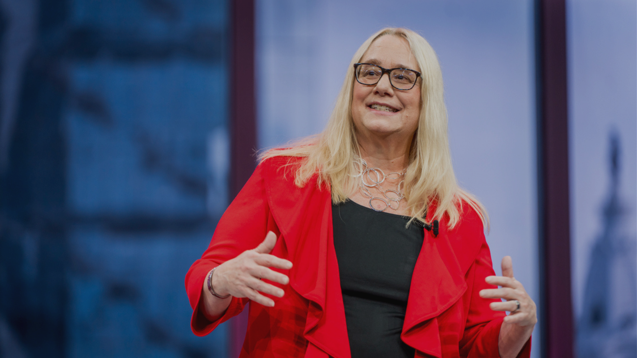 Dreama with long blonde hair wearing glasses and a red blazer speaking confidently on stage and gesturing during a presentation.