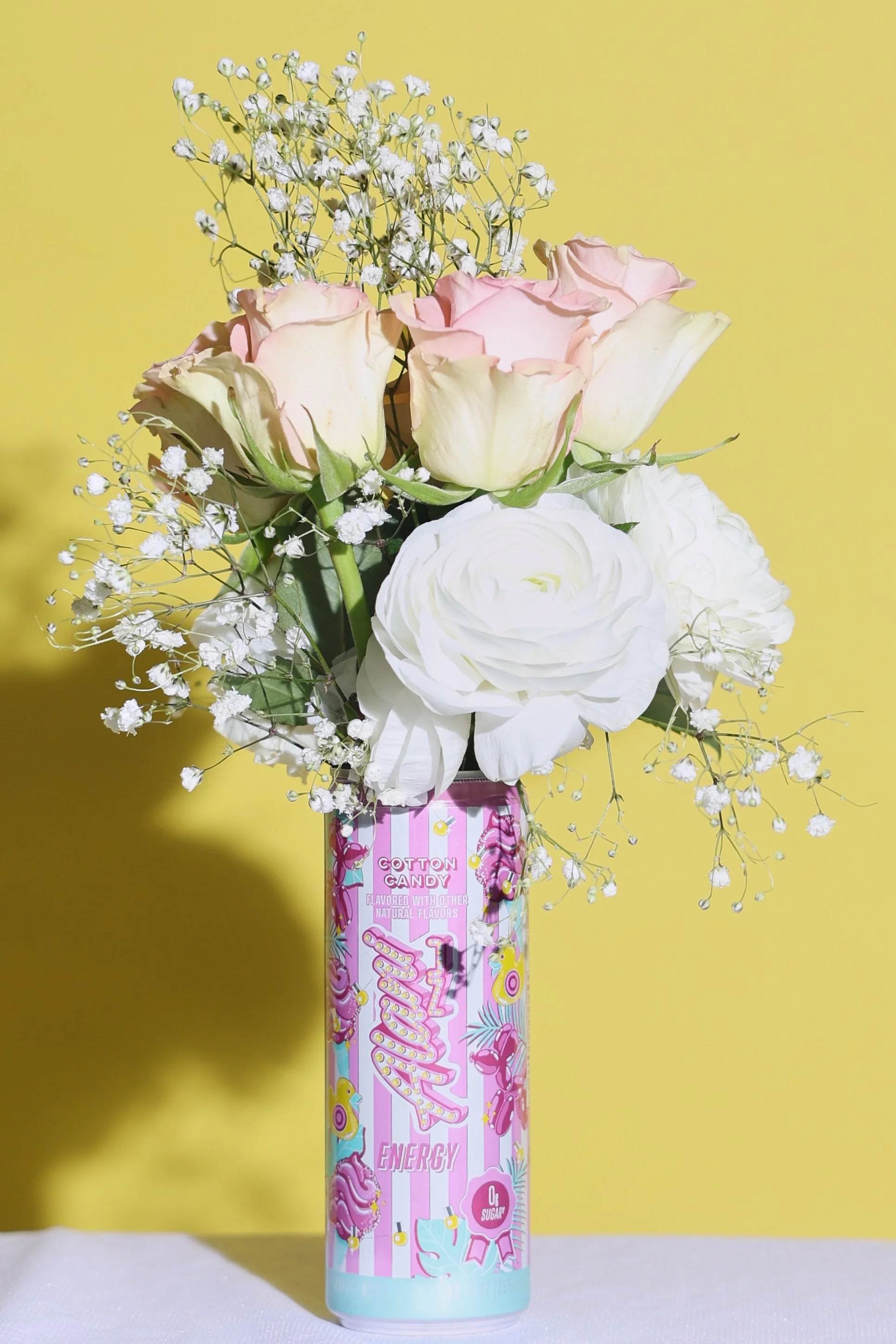 A colorful bouquet of pink and white roses with baby's breath in a pink and blue can labeled "Fruity Energy" with a yellow background.
