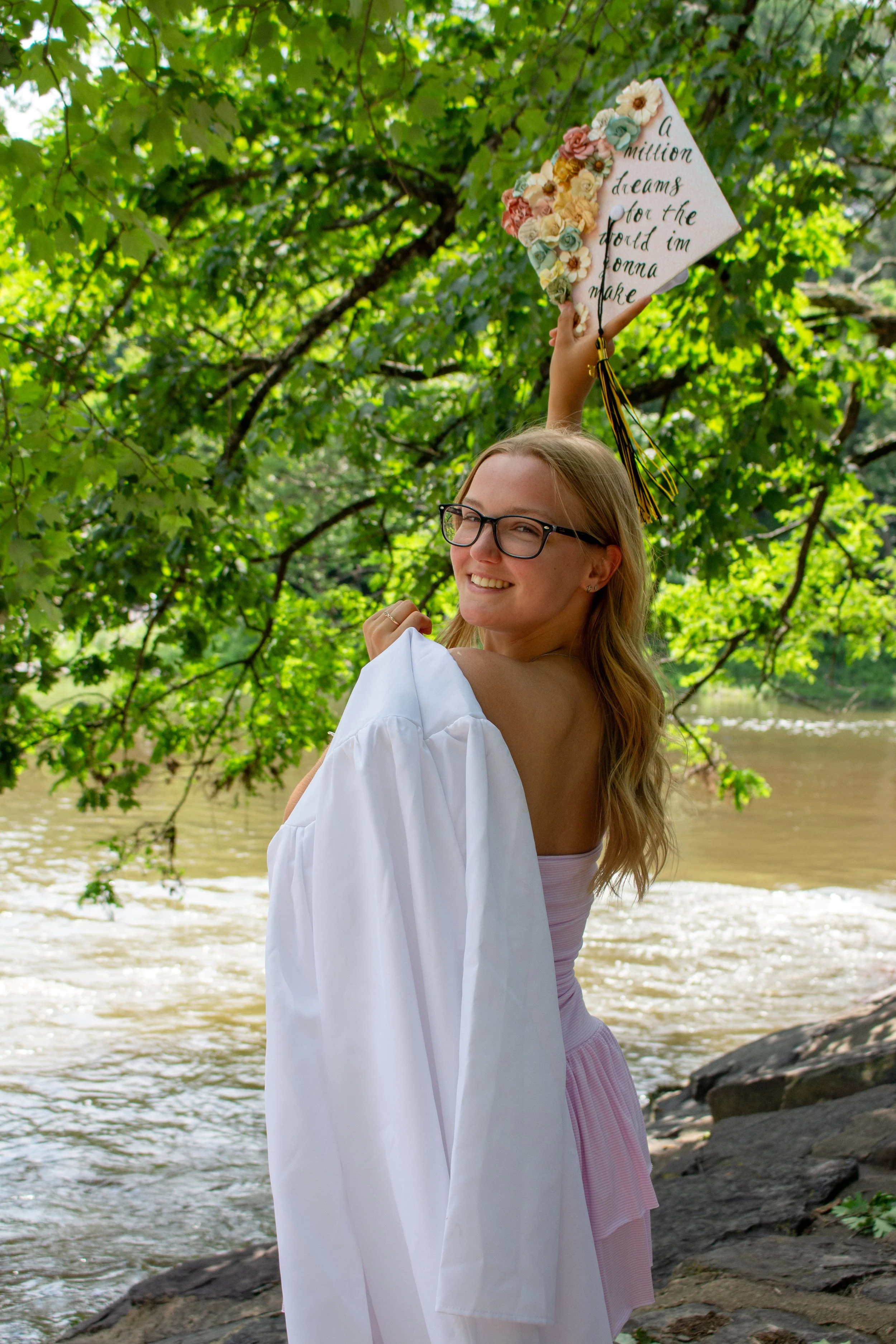 A woman with glasses and long brown hair smiling outdoors near a river, holding a graduation cap decorated with flowers and a quote, under green trees.