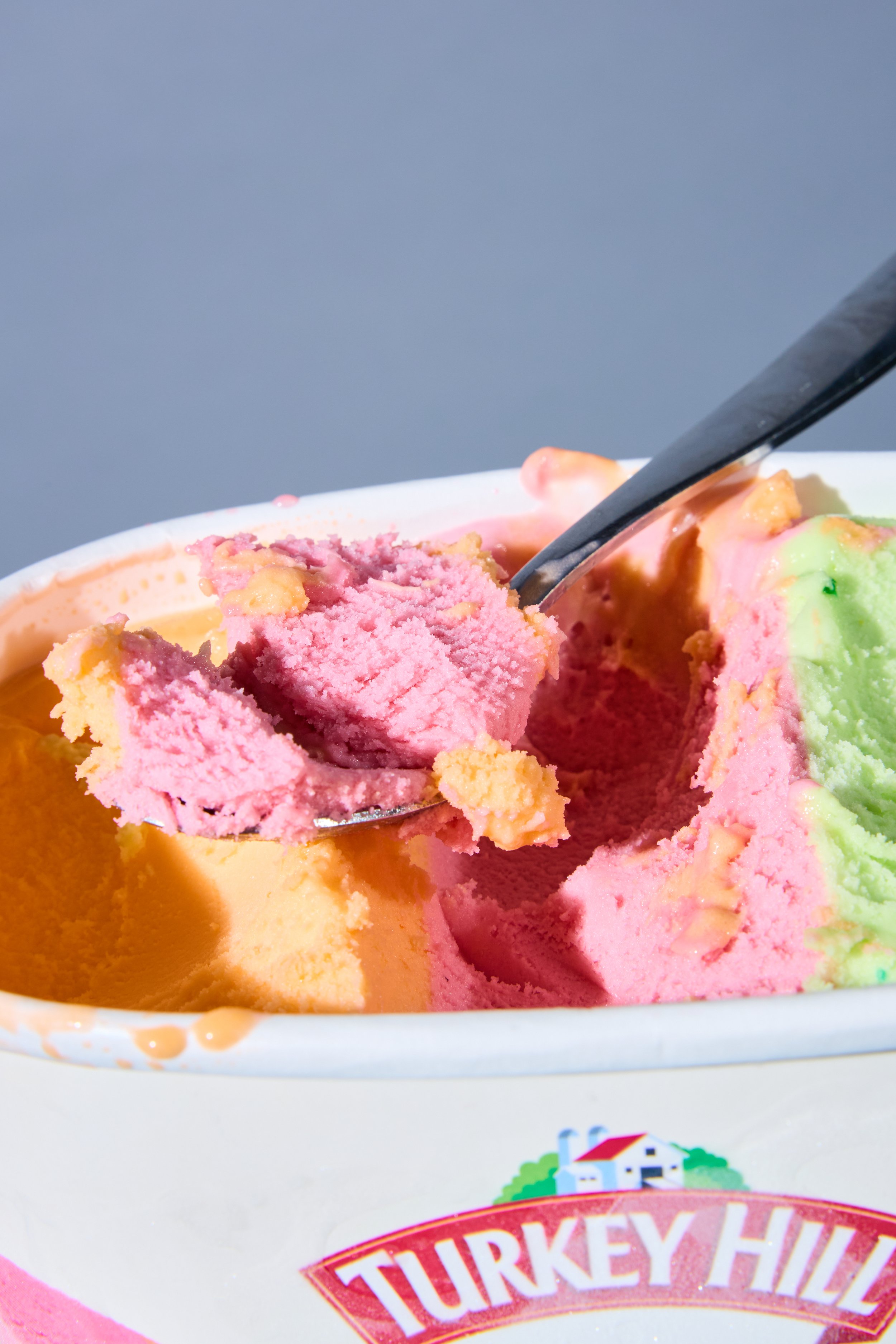 Close-up of a scoop of pink and green ice cream with a silver spoon in a white bowl, with part of a white ice cream container with a red and gold label reading "Turkey Hill" in the foreground.