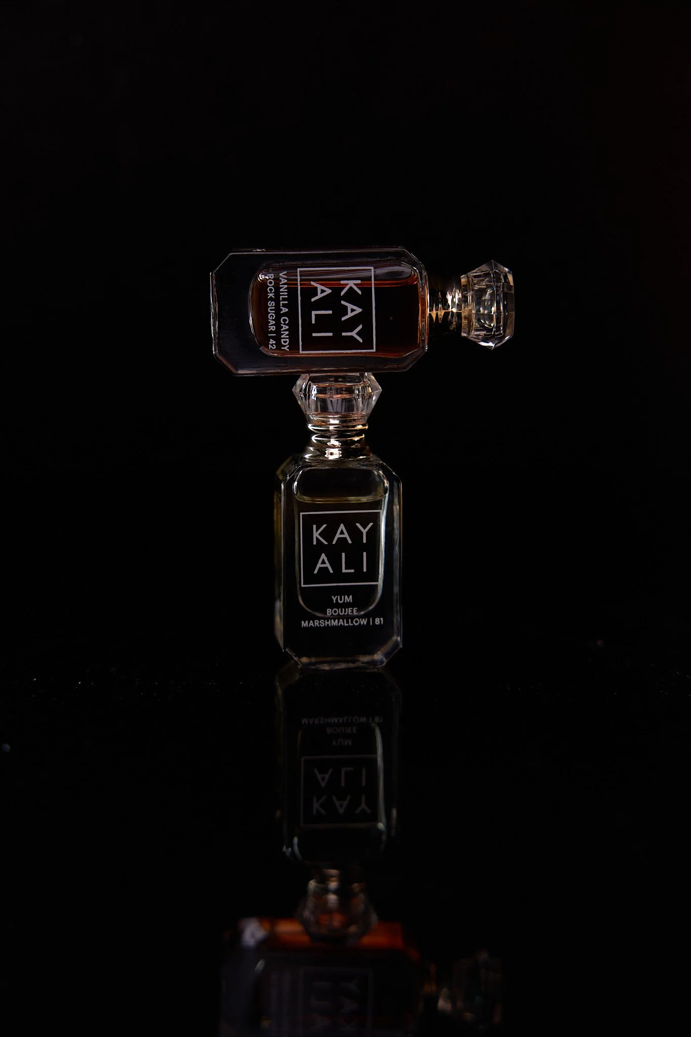 A glass perfume bottle labeled 'KAYALI' with a cap, placed on a reflective black surface, against a black background. The label indicates the scent is 'YUM' with marshmallow and 81.