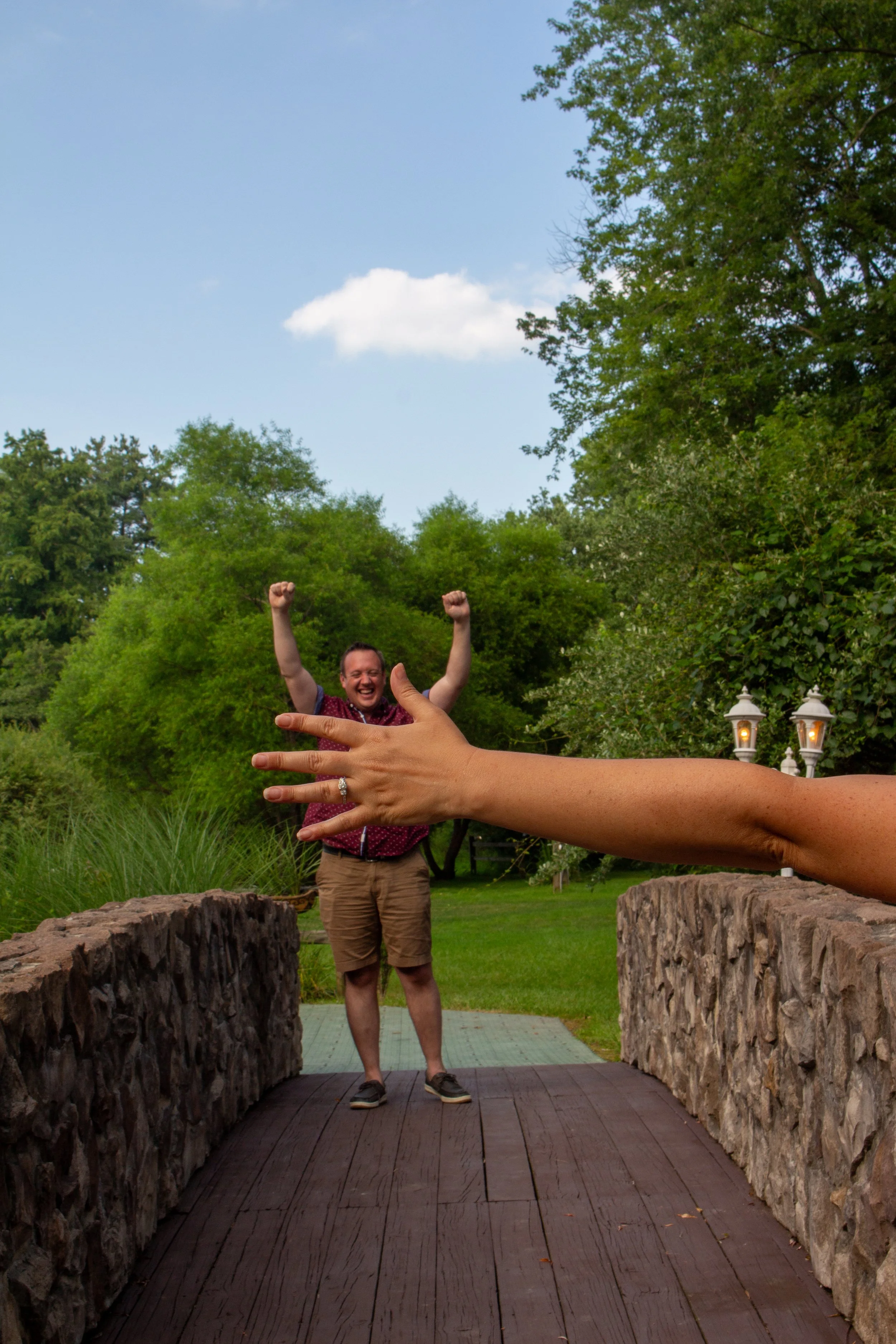 A smiling woman stands on a small stone bridge with wooden planks, surrounded by greenery, with a person behind her with raised arms and a blue sky with a few clouds overhead.