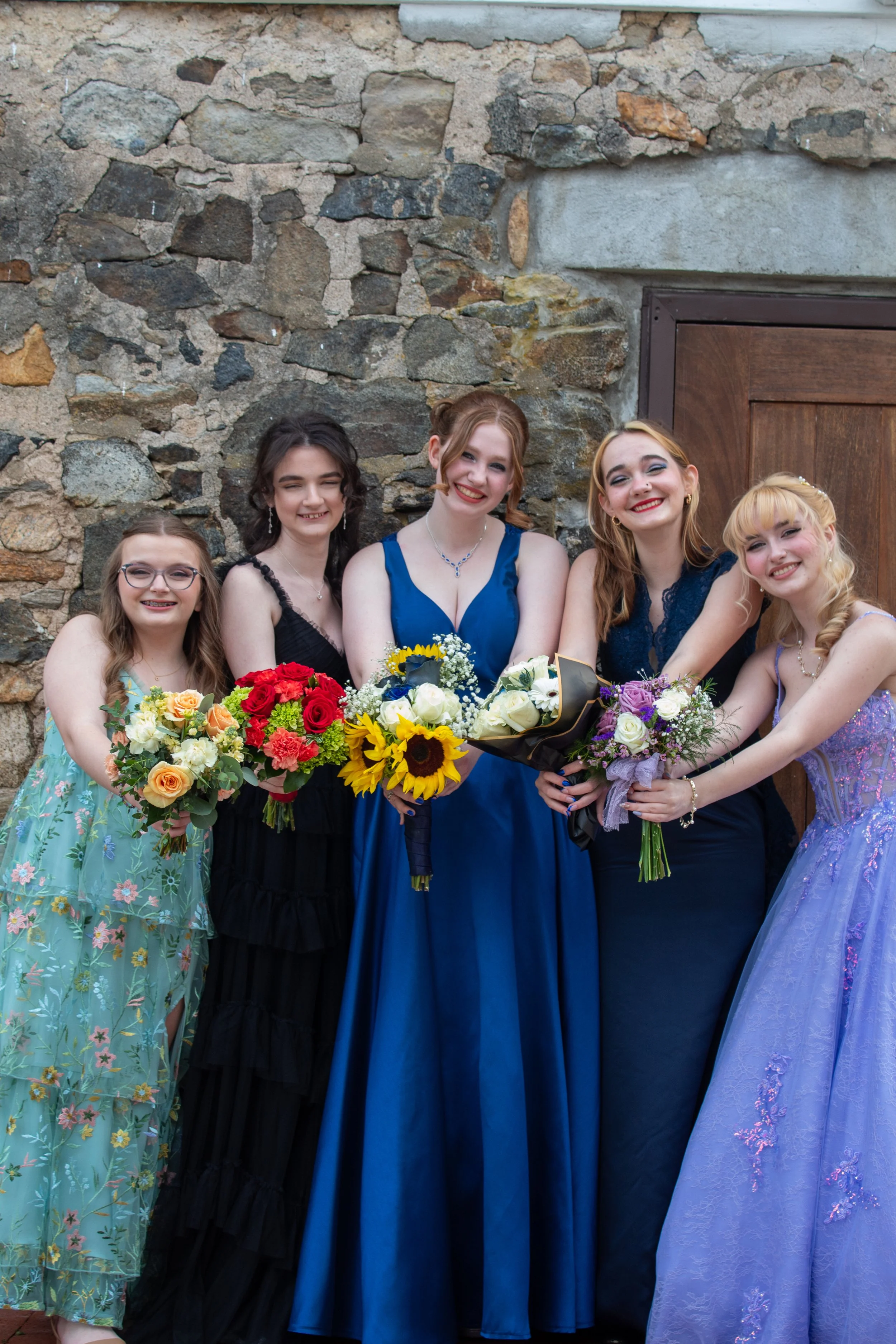 Group of five young women dressed in colorful formal prom gowns, holding bouquets of flowers, standing in front of a stone wall. They are smiling.