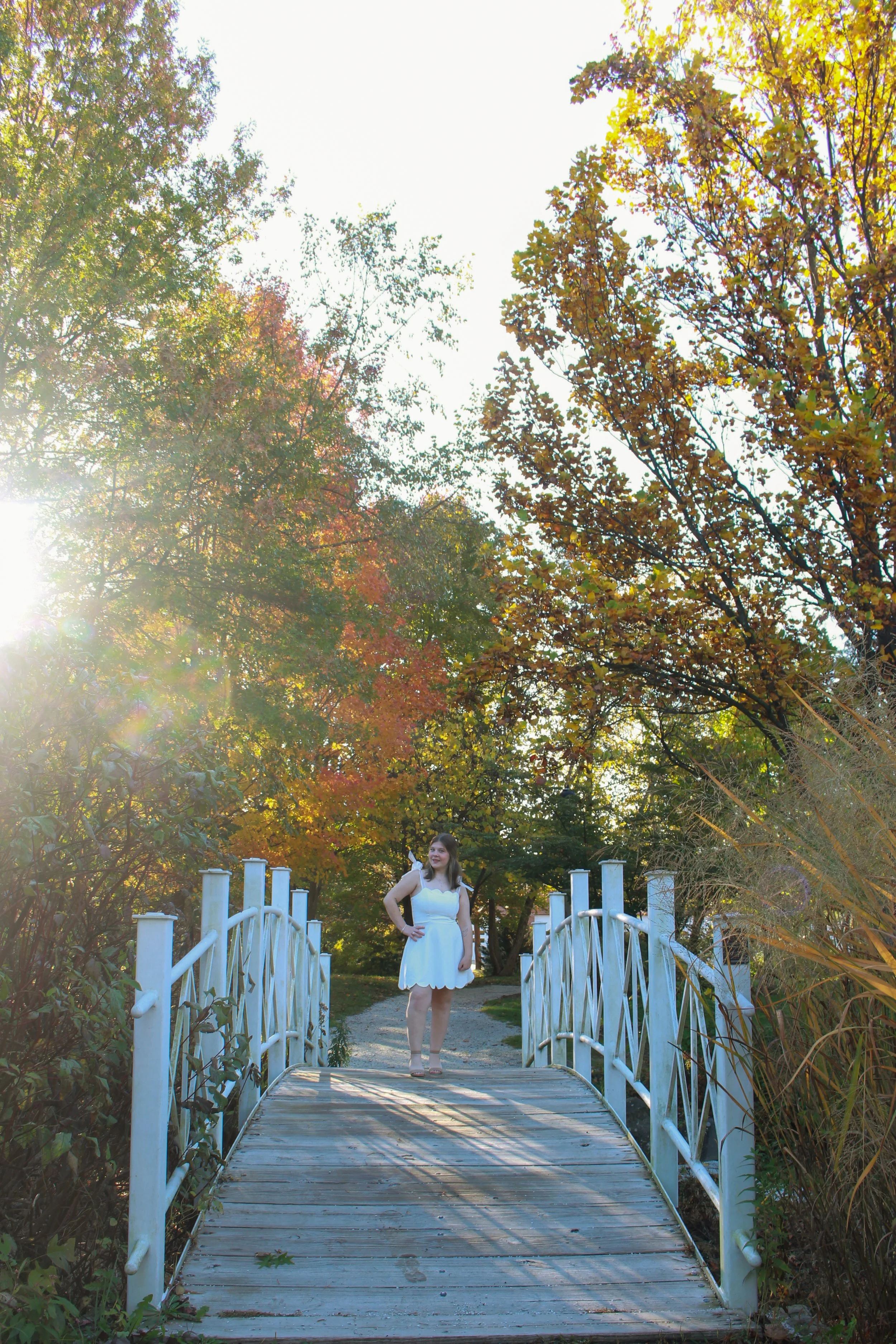 A woman in a white dress standing on a small bridge, surrounded by trees with fall foliage, sunlight shining through the leaves.