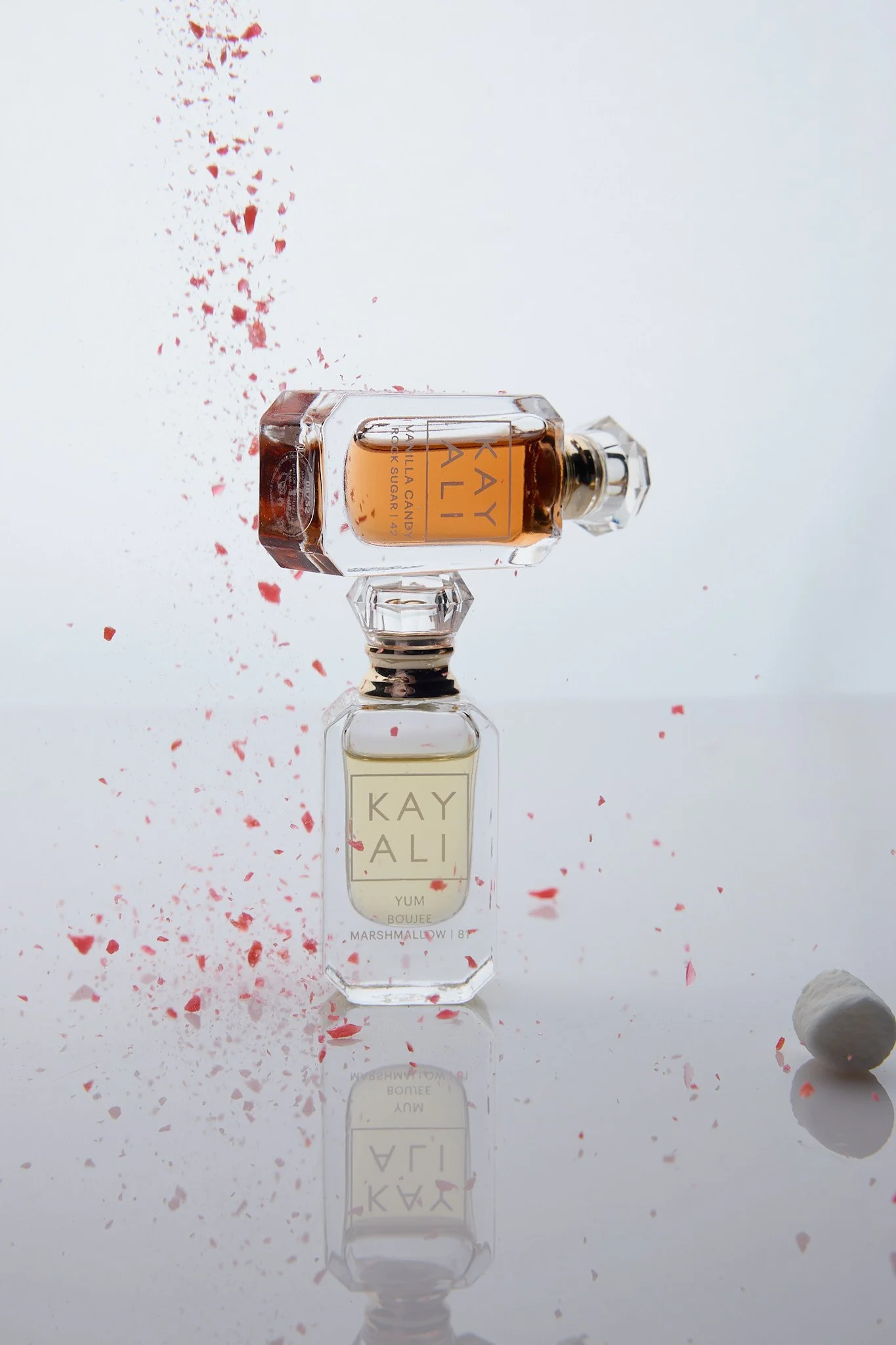 Two perfume bottles, labeled 'KAYALI,' are positioned on a reflective surface with red and white confetti scattered around. One bottle is upright, and the other is tilted, with the foam or confetti falling from the tilted bottle.