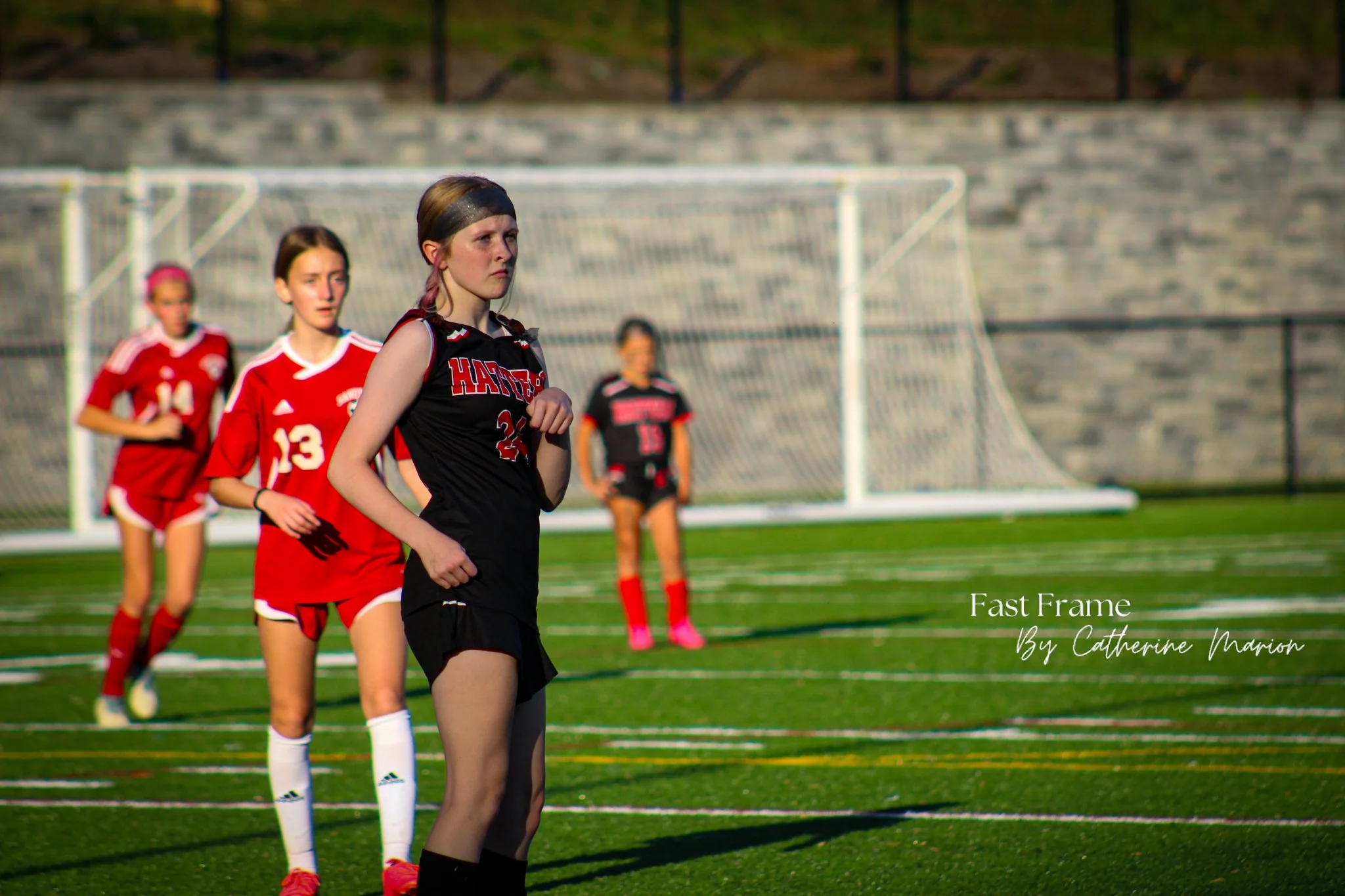 A group of young female soccer players standing on a soccer field during a game or practice. They are wearing sports uniforms with some in red and one in black, with a goalpost in the background. The sunlight suggests it is late afternoon or early ev
