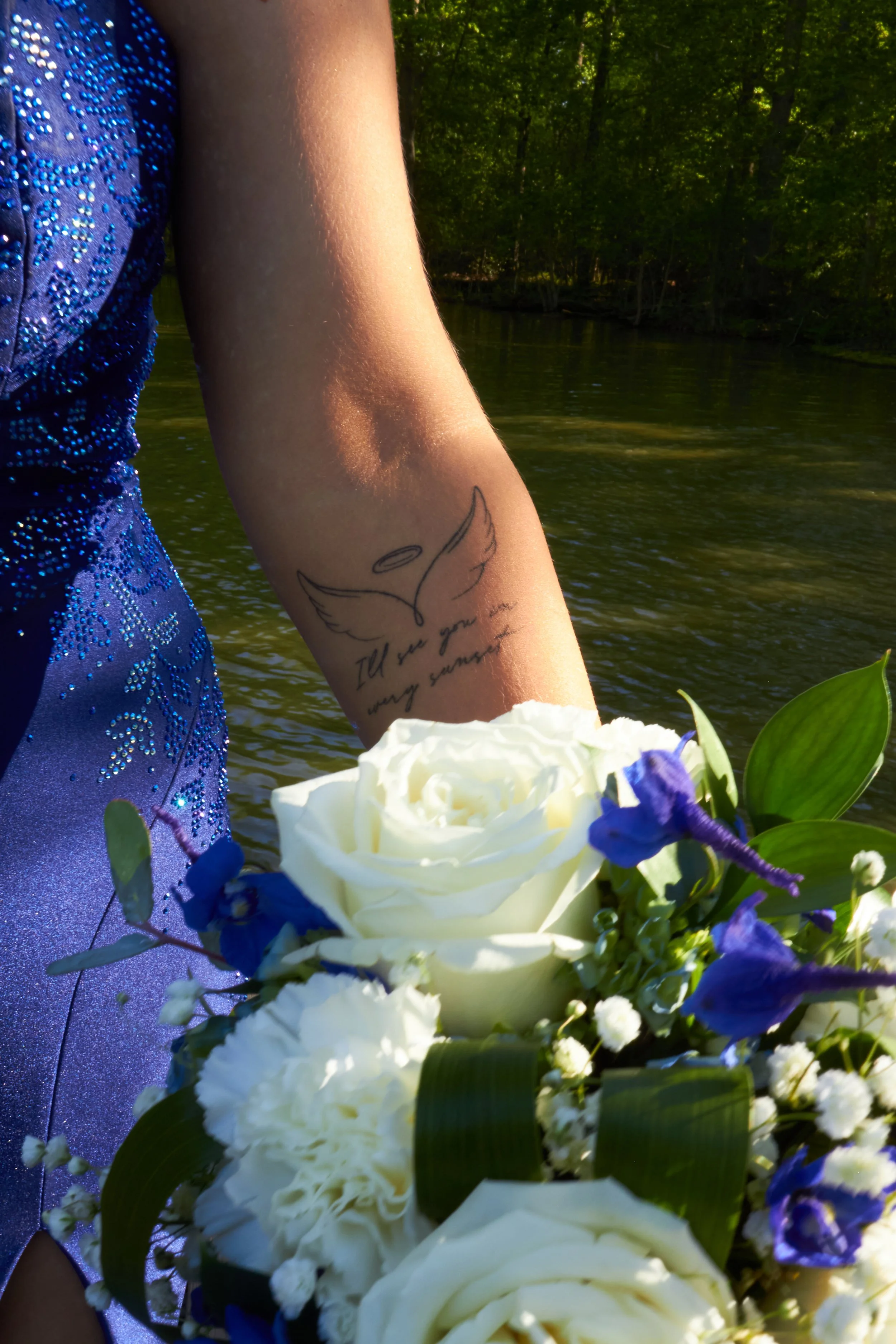A person's arm with a tattoo of angel wings and a halo, holding a bouquet of white roses and purple and white flowers near a river with green trees in the background.