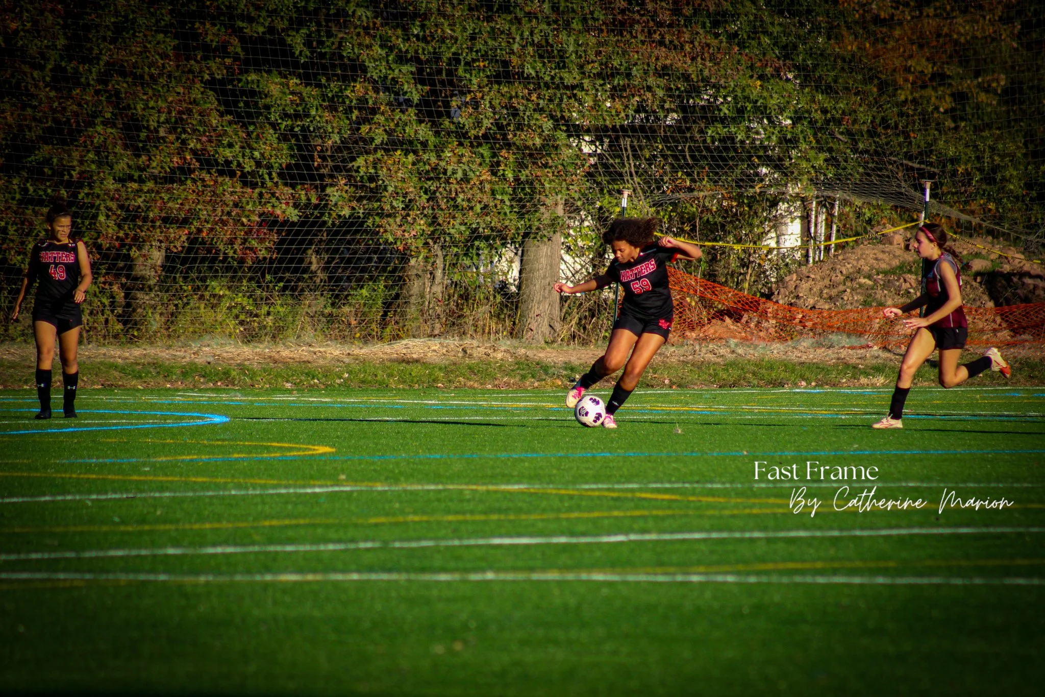 Three young female soccer players in black and red uniforms practicing on a green field with white, blue, and yellow lines, with trees and netting in the background.