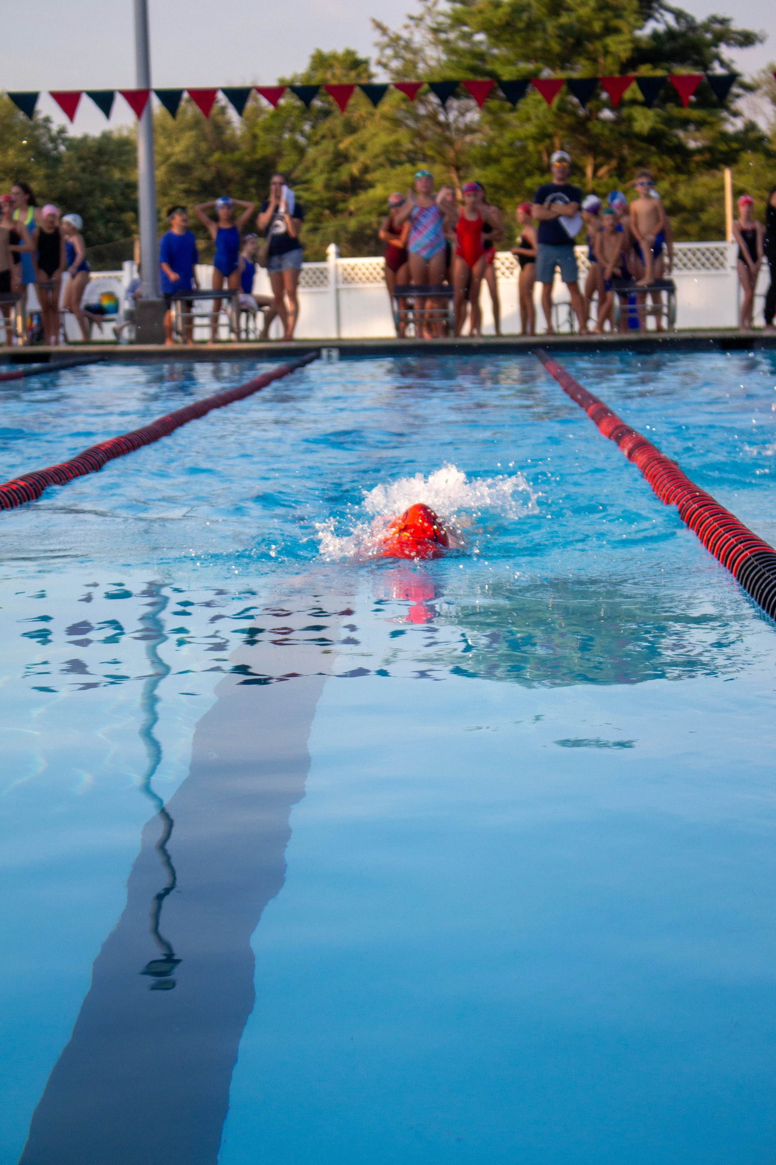 Swimmer in orange cap glides through a swimming pool, with a group of people, likely spectators or coaches, watching from the pool deck in the background.