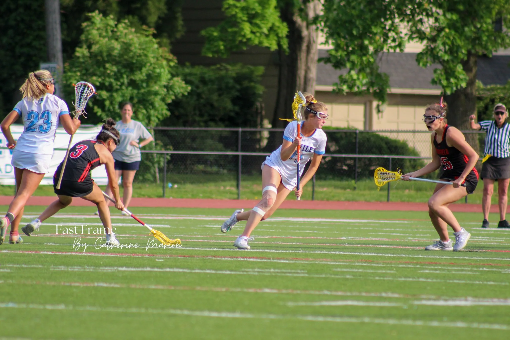 Girls playing lacrosse on a green field with trees in the background, wearing protective goggles, holding lacrosse sticks, and dressed in sports uniforms.