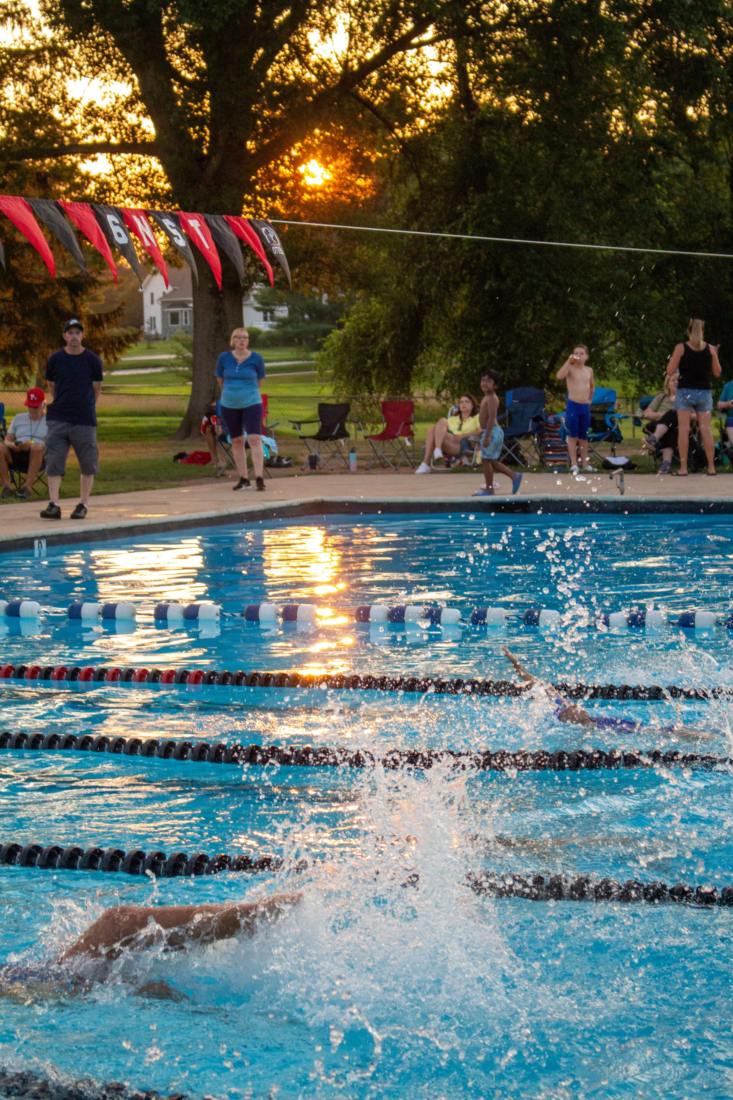 A group of people, including children and adults, watching a swimming competition at an outdoor pool during sunset, with the swimmer in the water, splashing and racing in the lane. There are chairs and spectators on the pool deck, and a large tree wi
