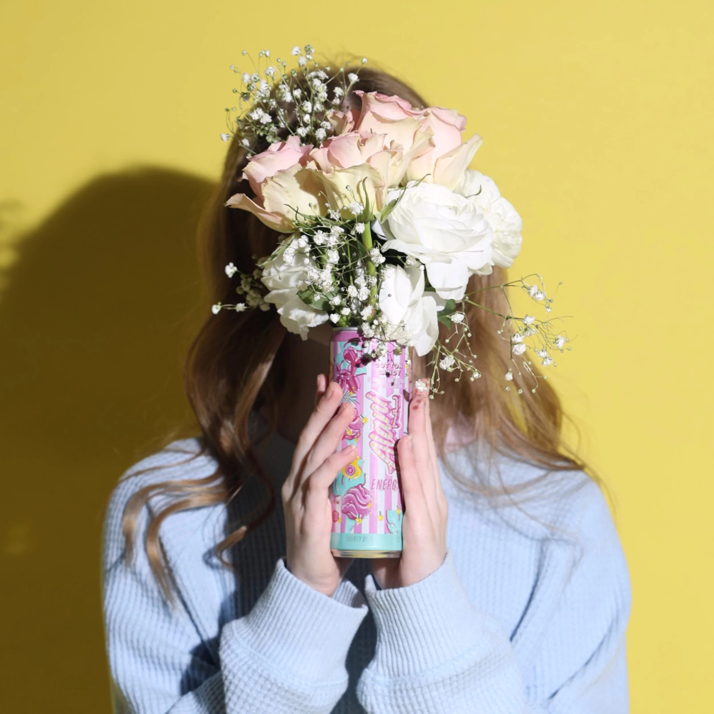 Person holding a colorful can of cotton candy-flavored energy drink with a large bouquet of white and pink flowers obscuring their face, against a bright yellow background.