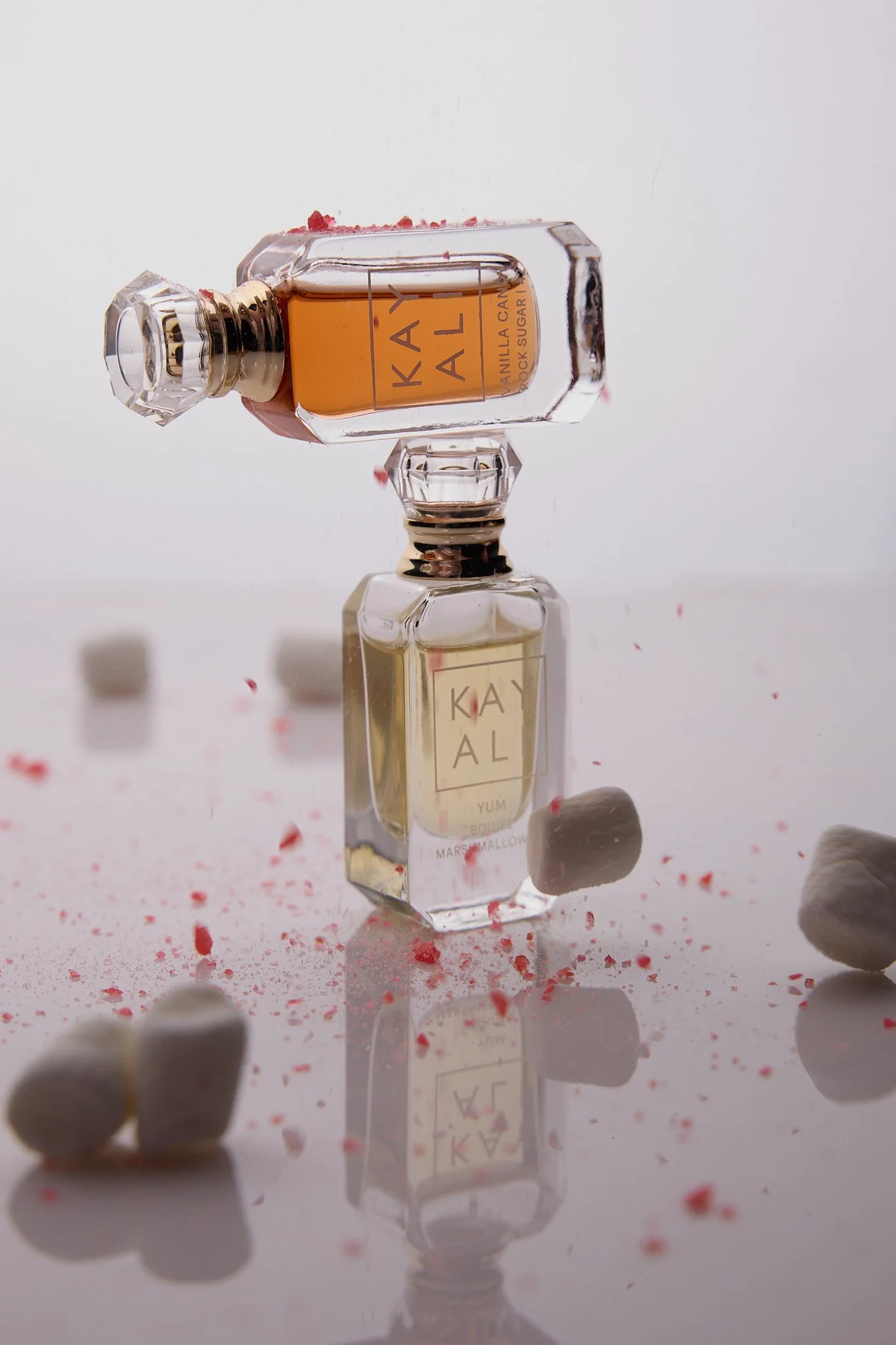 A small glass perfume bottle lying on its side with pink liquid inside, surrounded by scattered pink and white pills on a reflective surface.