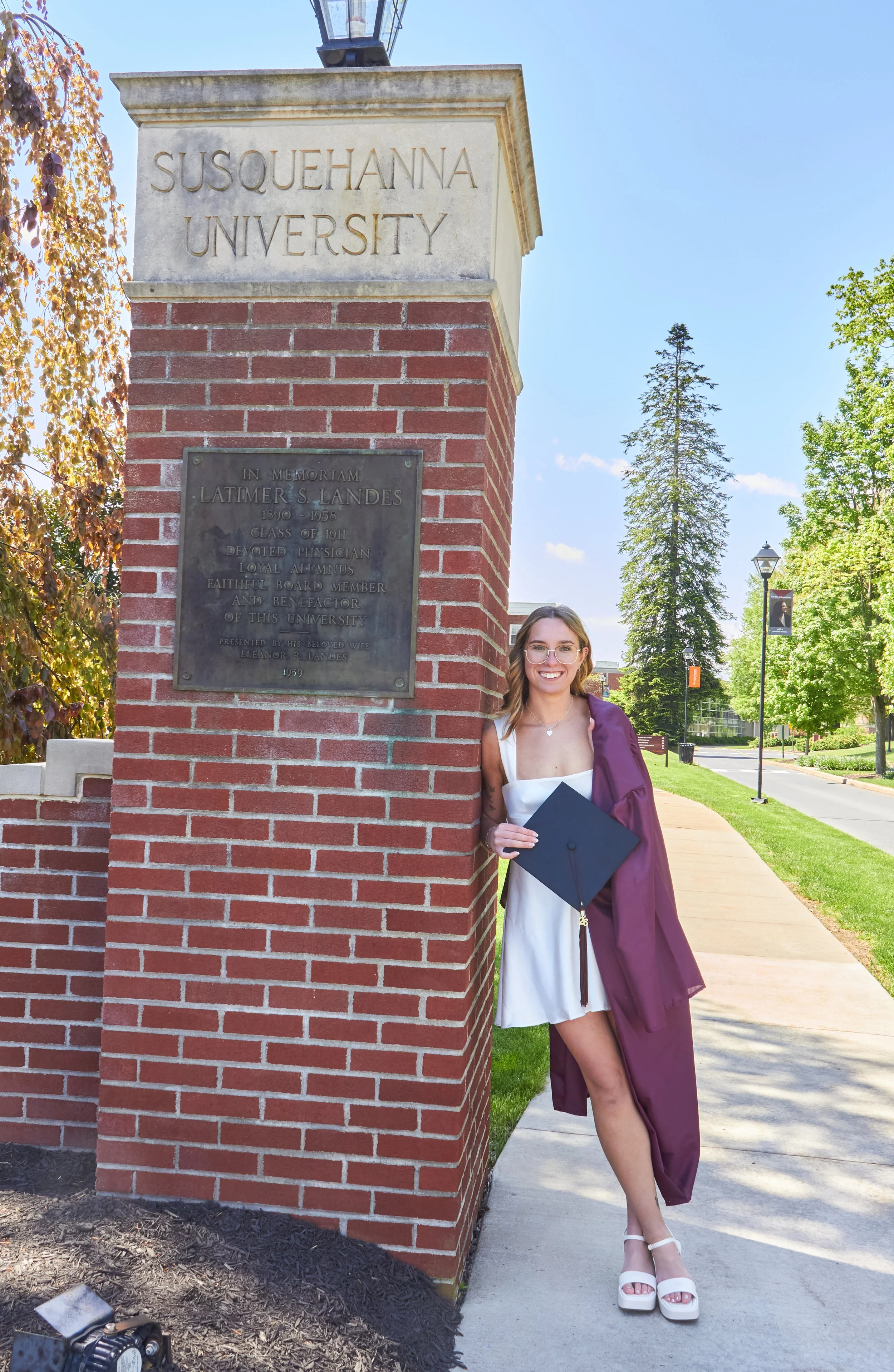 A young woman in a white dress and white platform shoes, wearing glasses, and holding a graduation cap, stands next to a brick pillar with a university sign. The sign reads 'Susquehanna University' and has a plaque in memory of Latimer S. Landes. The