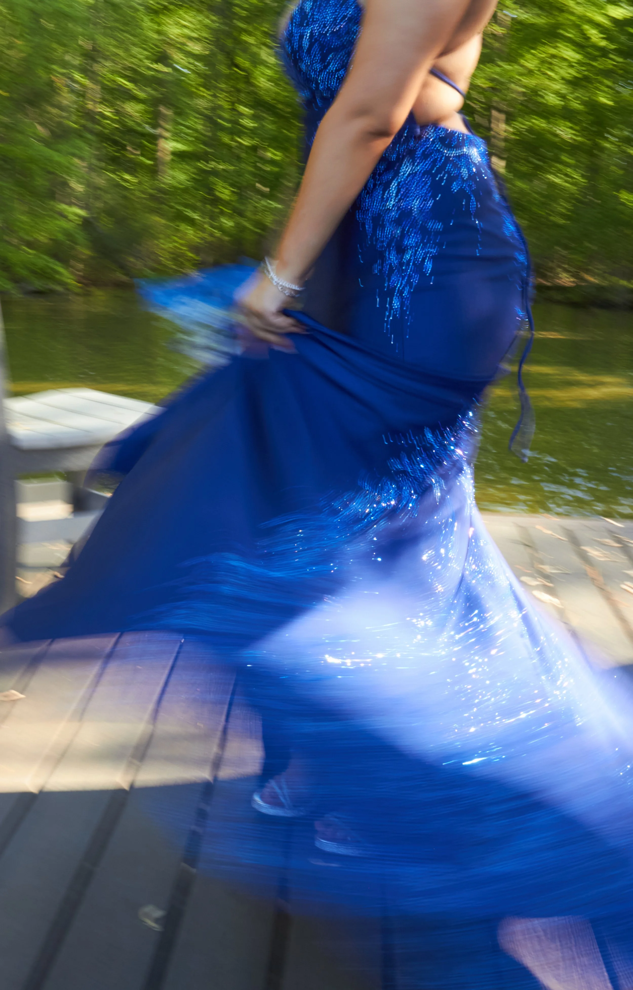 A woman in a shiny blue dress walking on a wooden dock near a body of water surrounded by green trees.