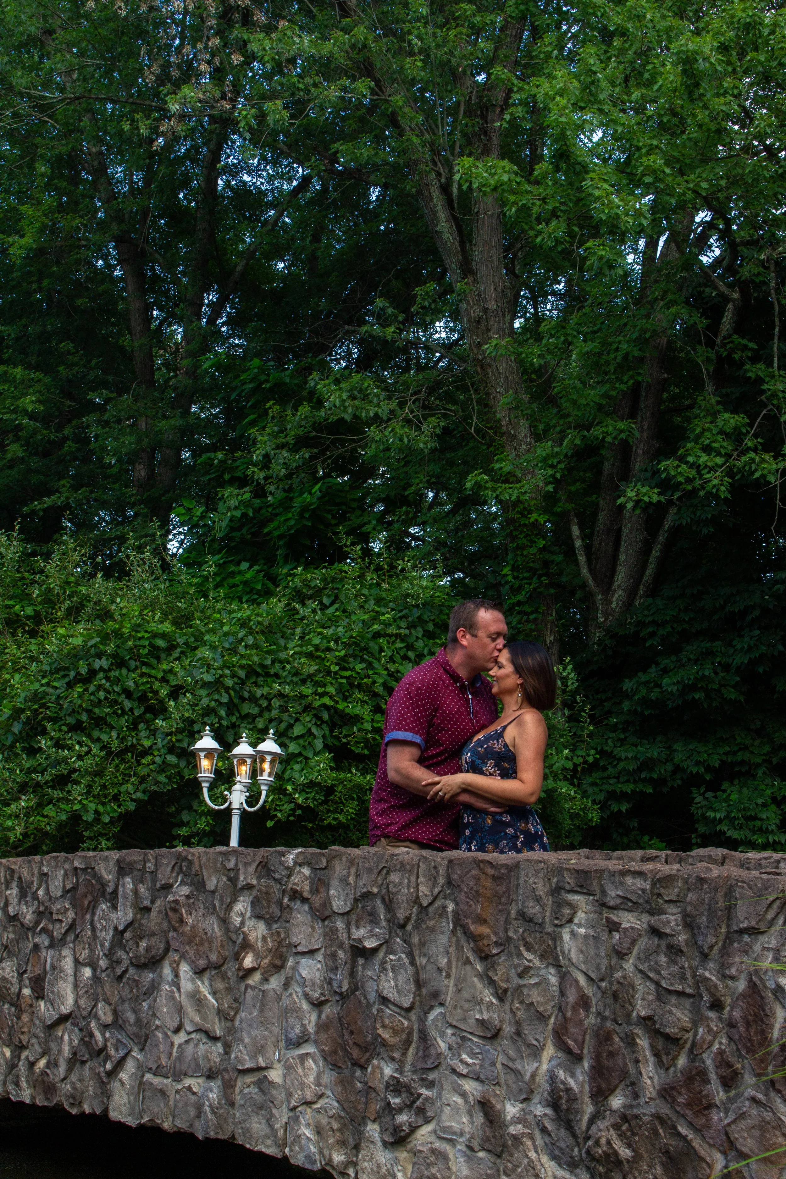 A man and woman standing close together on a stone wall, kissing each other, with lush green trees in the background and a white lamp post nearby.