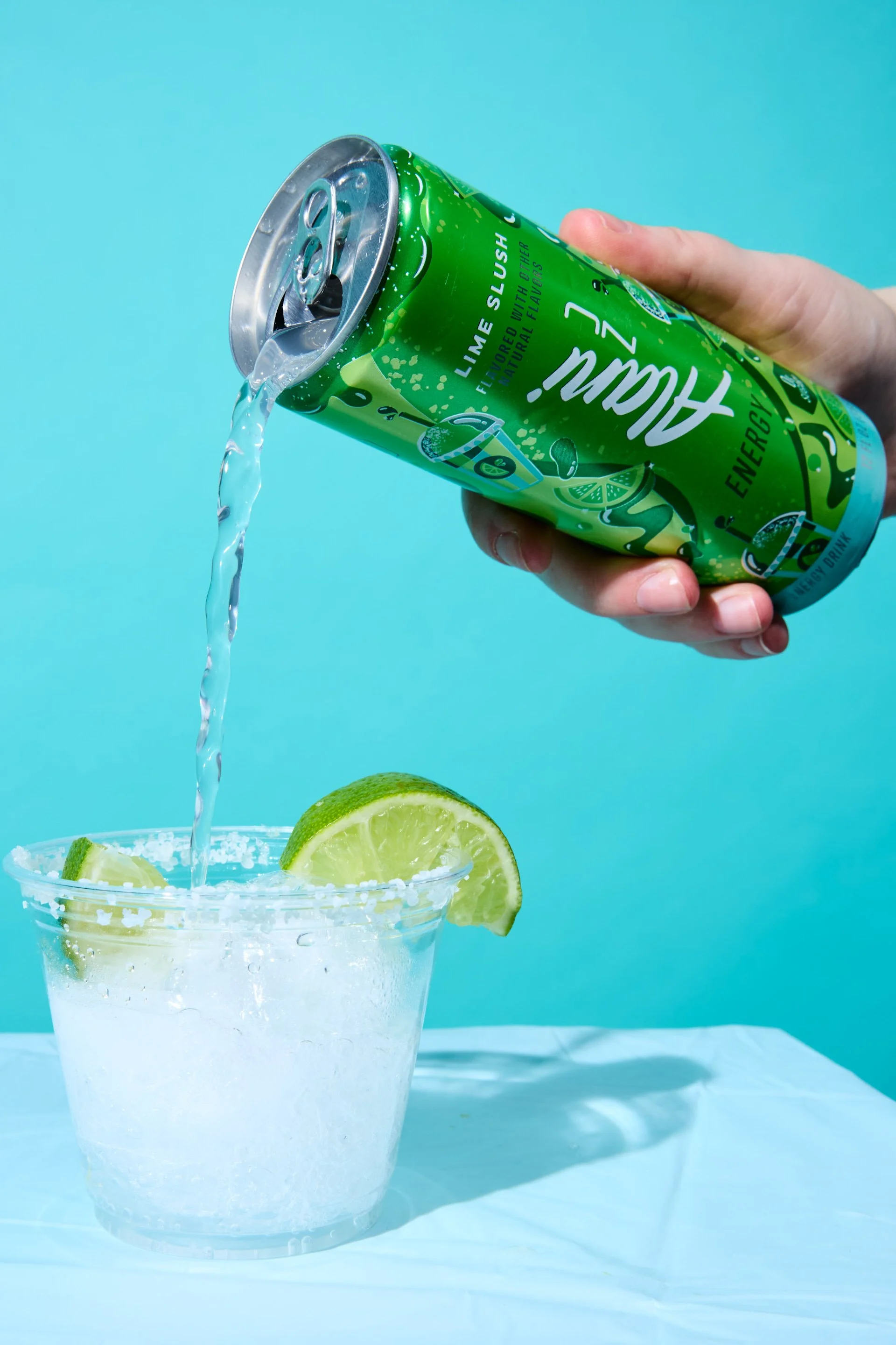 A hand pouring lime-flavored energy drink from a can into a glass with crushed ice and lime wedges on the rim, against a blue background.