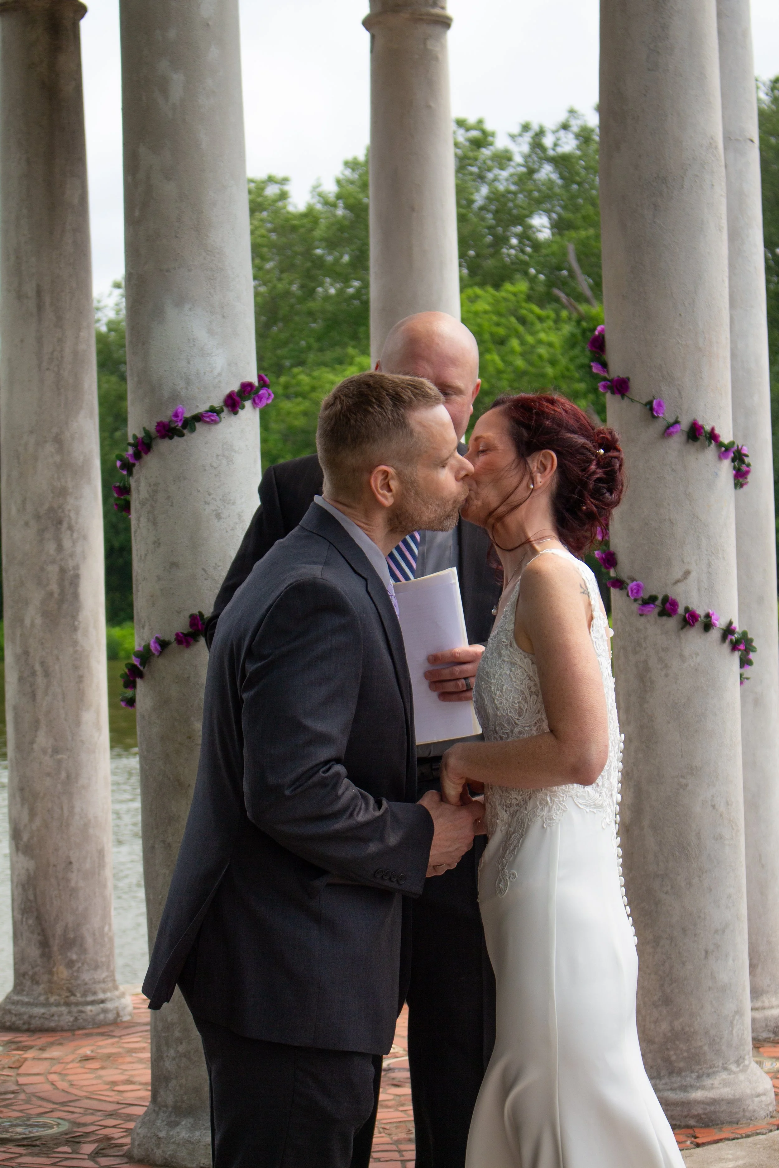 A couple in wedding attire kissing during their wedding ceremony, with a man reading vows behind them, in front of stone columns decorated with purple flowers, by the water with green trees in the background.