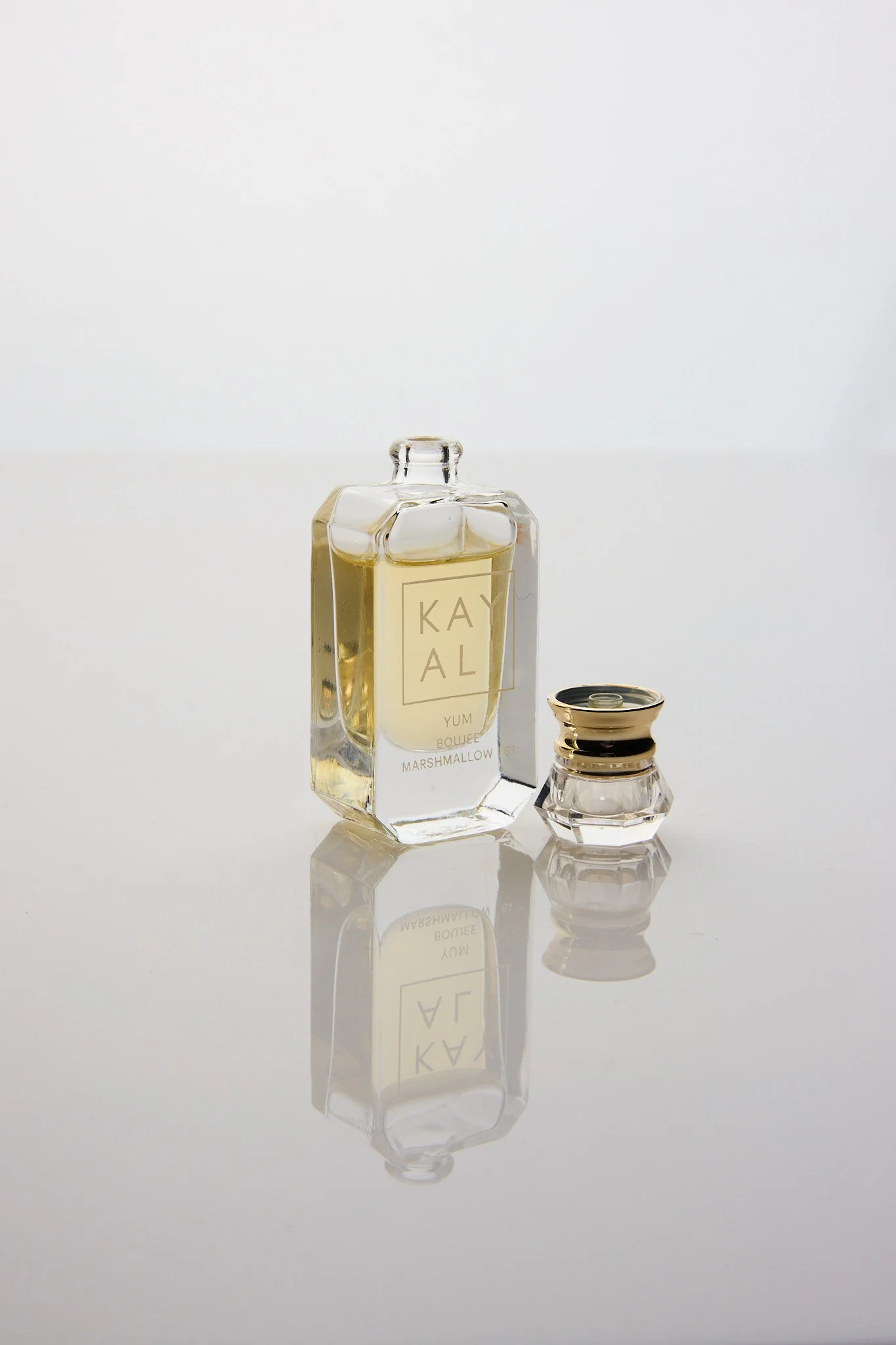 A clear glass bottle with golden liquid labeled "KAY AL" and a smaller glass container with a reflective metal lid, both placed on a reflective surface against a plain white background.