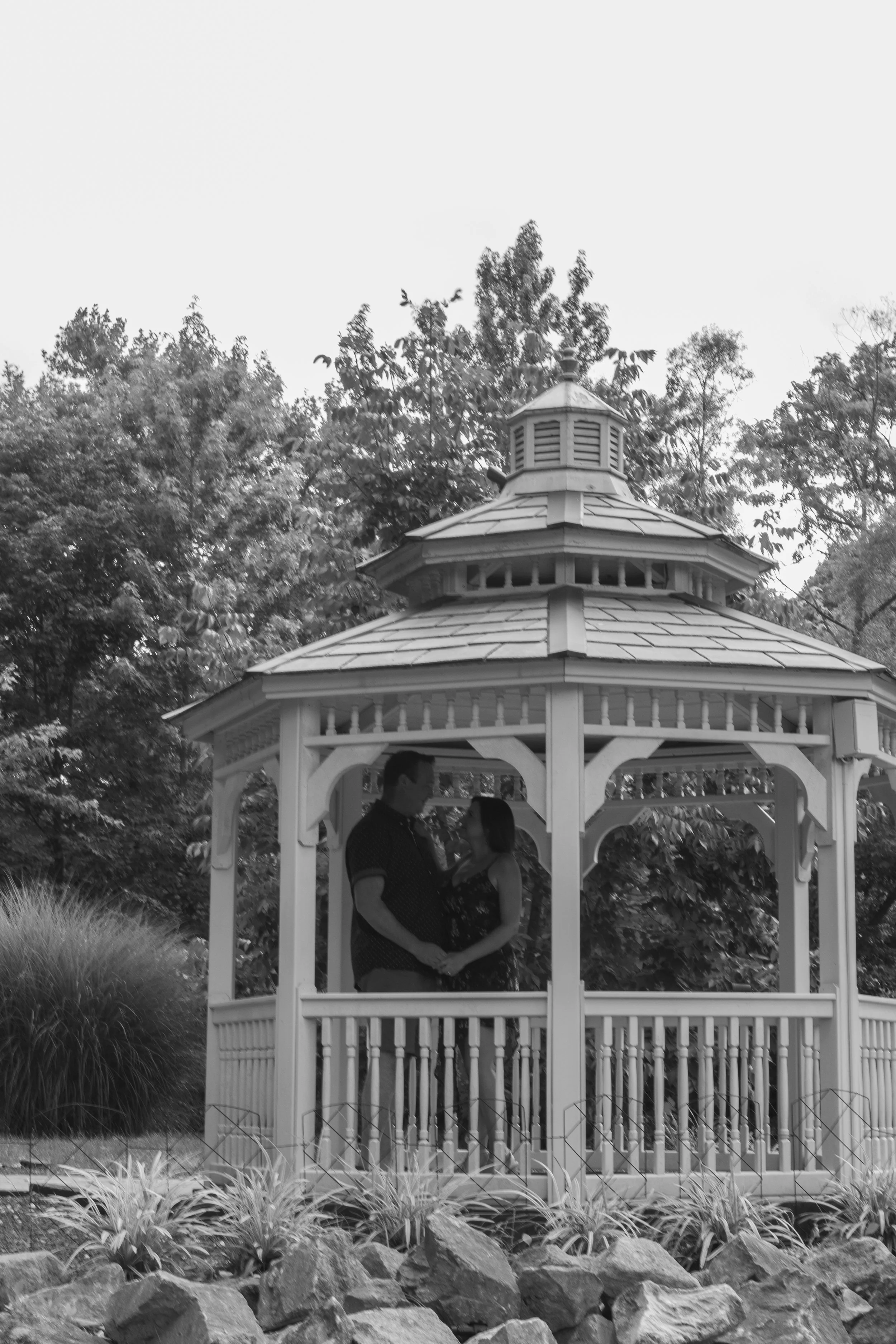 A couple standing closely together in a gazebo, surrounded by trees and plants, with rocks in the foreground.