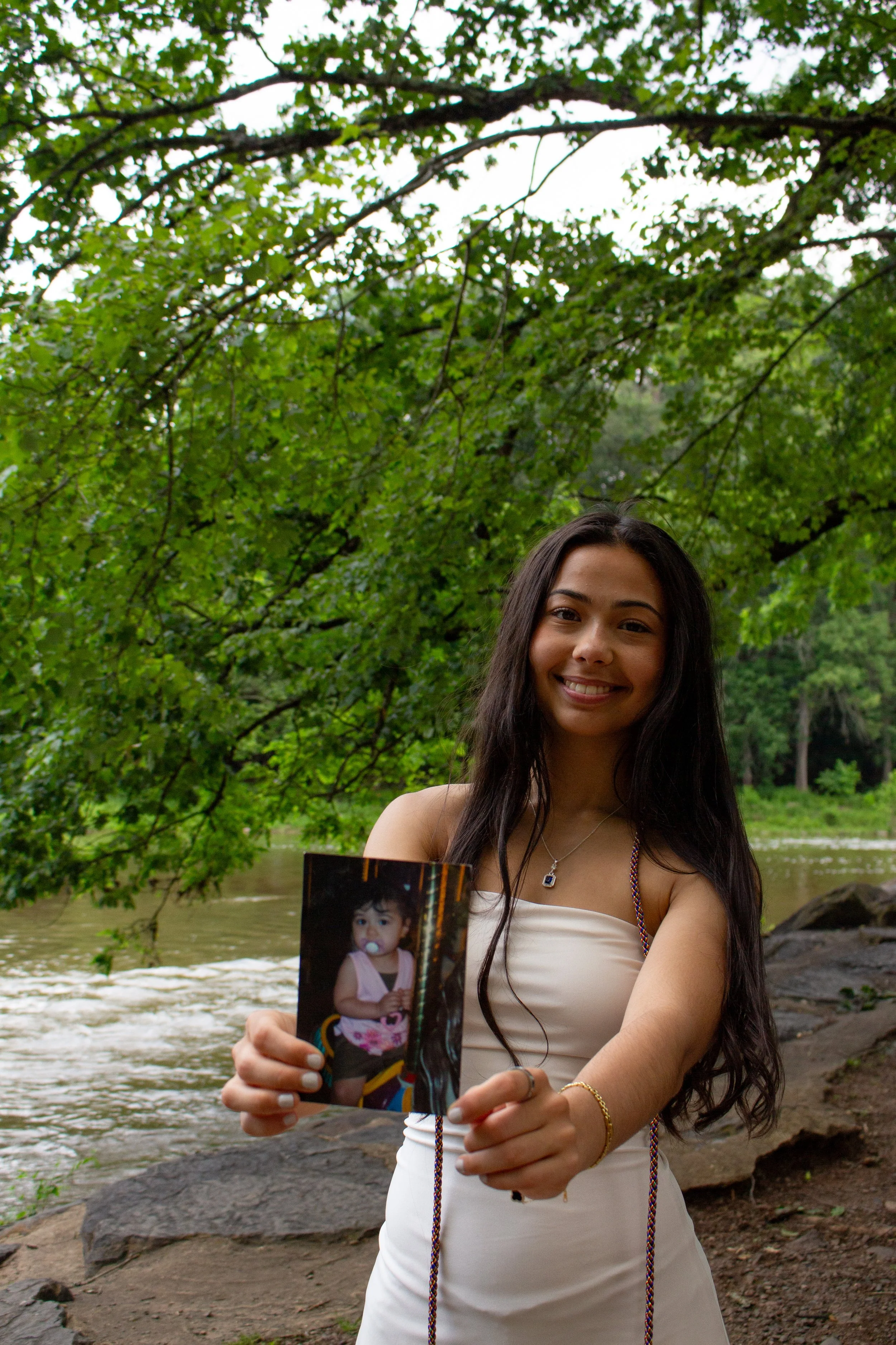 A young woman with long dark hair smiling and holding a photo of a young girl with a pacifier, in an outdoor setting near a river and trees.