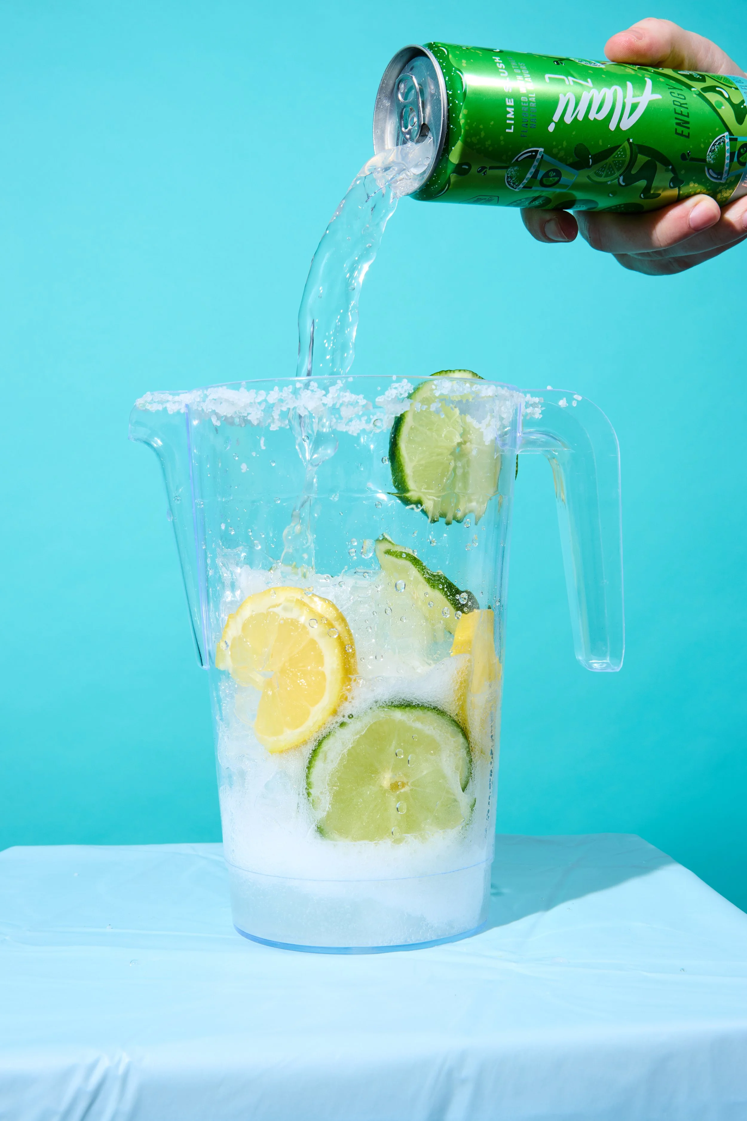 A hand holding a green lime-flavored energy drink can pouring into a clear pitcher with lemon and lime slices on a light blue background.