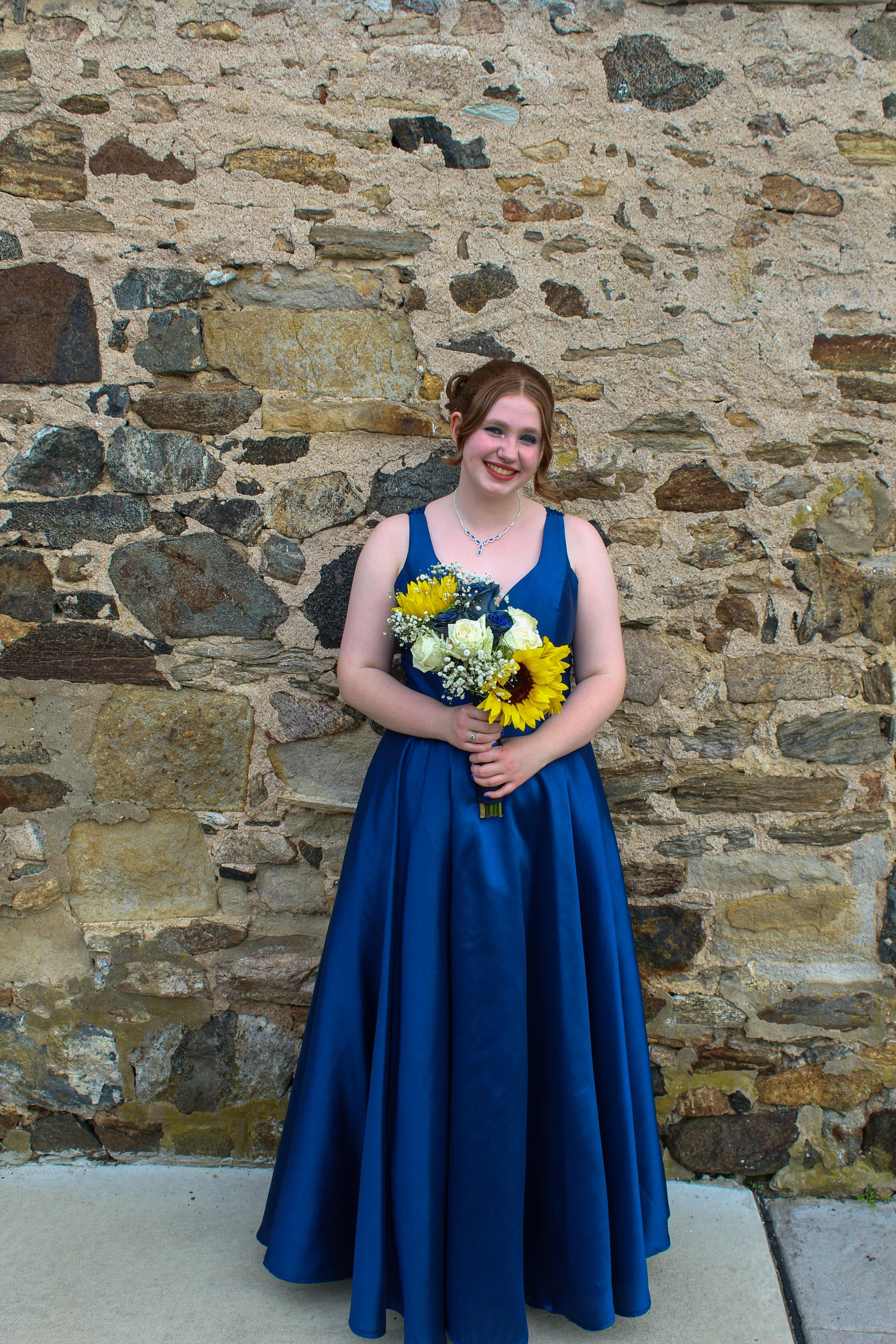 A young woman in a blue formal dress holding a bouquet of yellow and white flowers, standing in front of a stone wall.