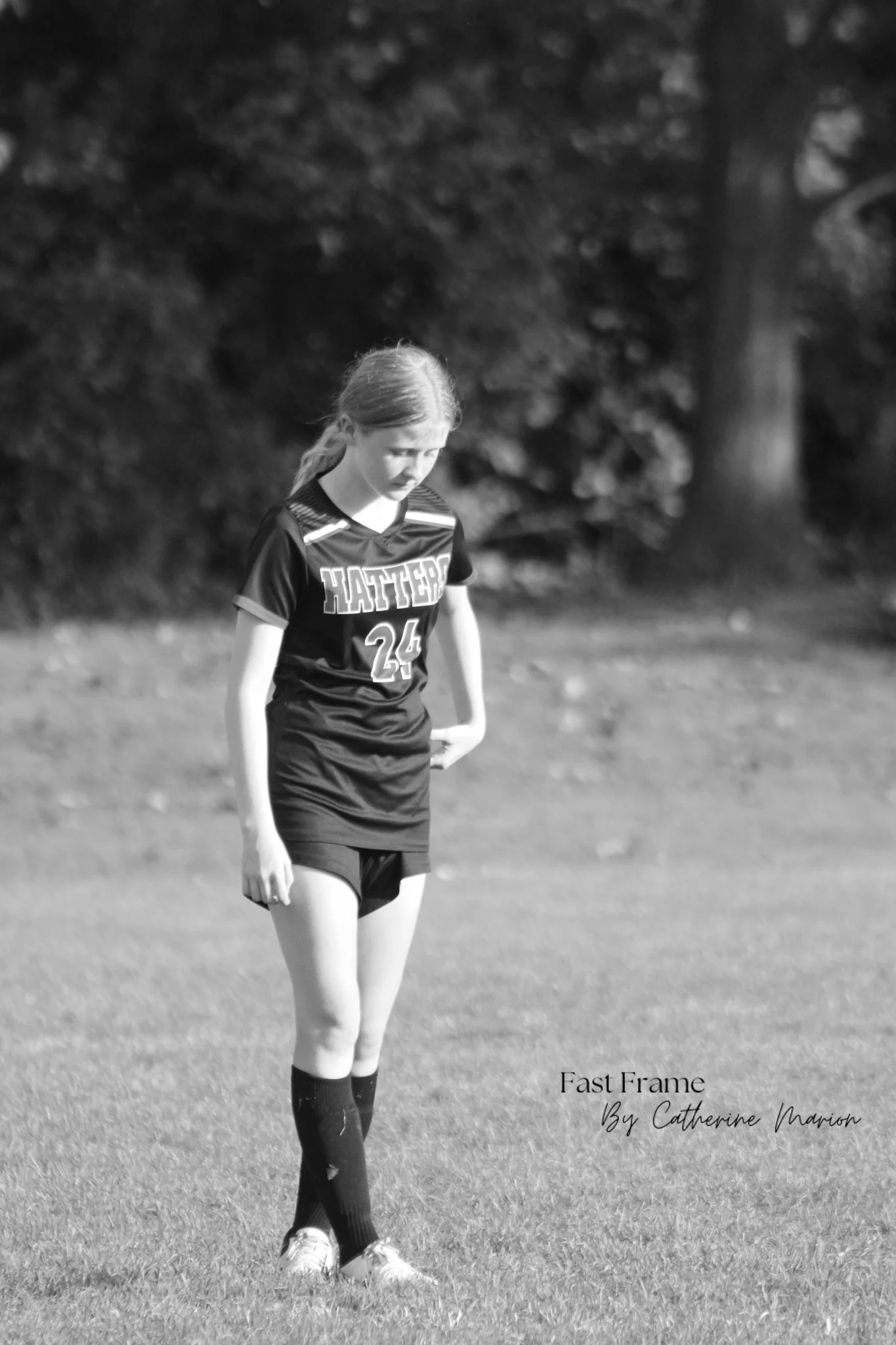 A young female soccer player standing on a grassy field, wearing a sports uniform with the number 24, looking down, with trees blurred in the background.