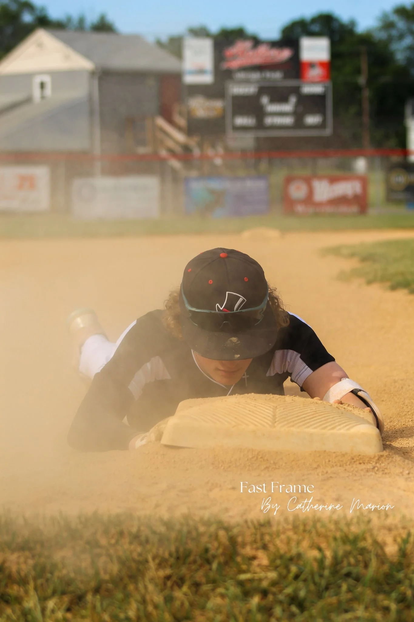 A person lying on a dirt track at a sports field with their face down near a starting block, wearing a cap. The background shows signs, a building, and trees out of focus.