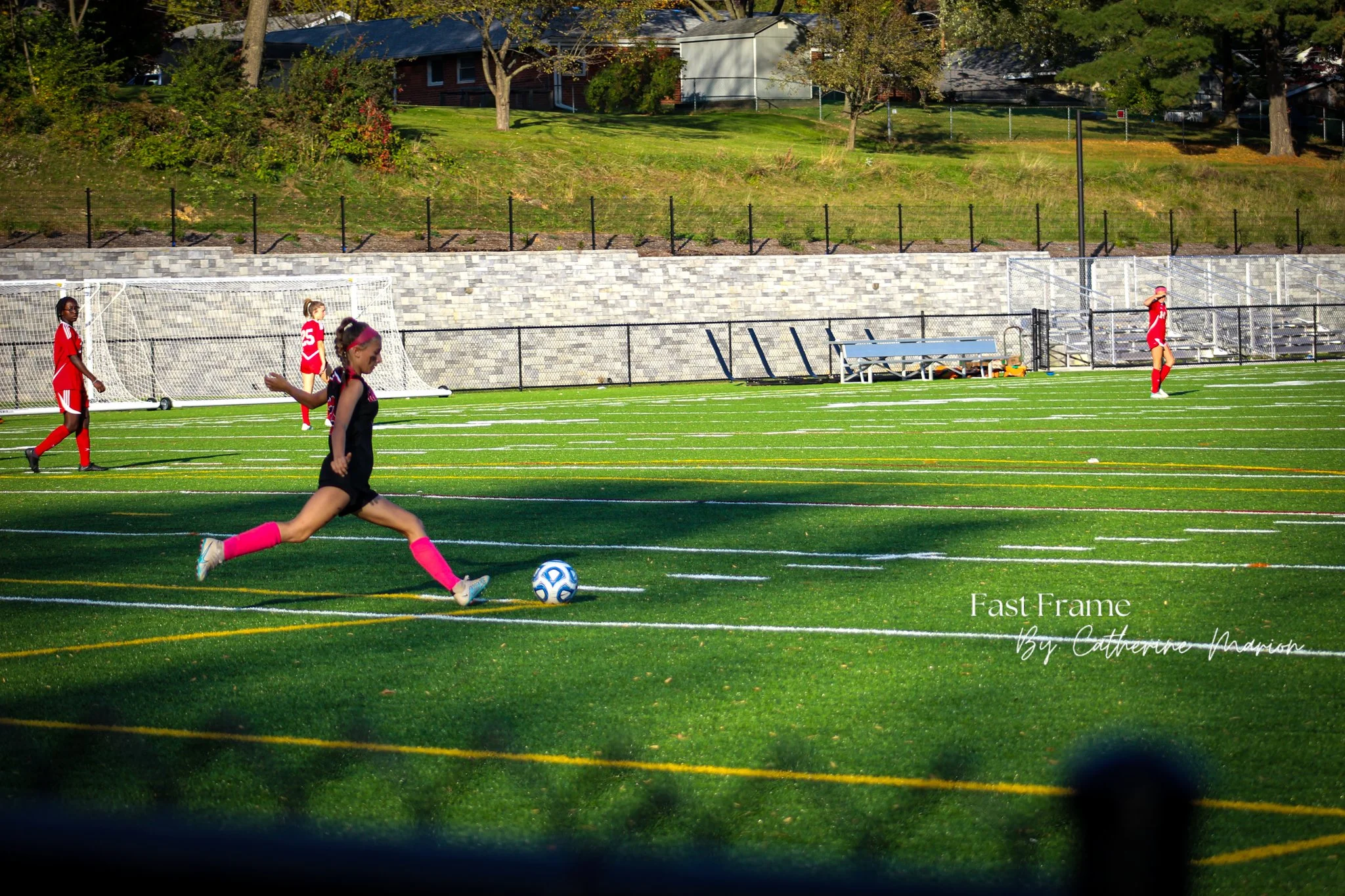 A young girl dressed in a black and pink outfit is running on a green artificial turf sports field, kicking a blue and white soccer ball. In the background, other children in red sports uniforms are standing near soccer goals, with trees, a brick bui