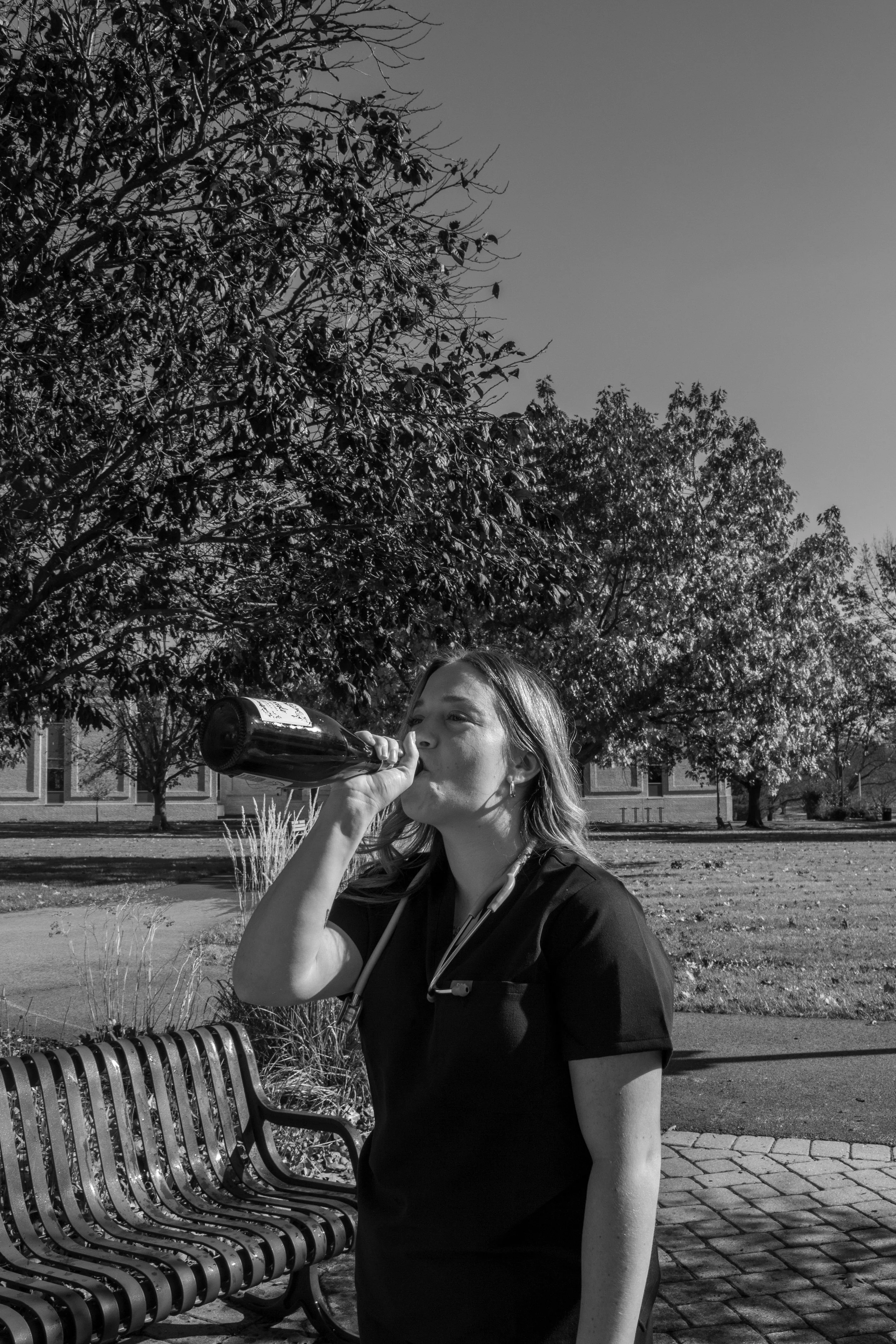 A woman in a black medical uniform drinking from a bottle, sitting on a park bench with trees and a building in the background.
