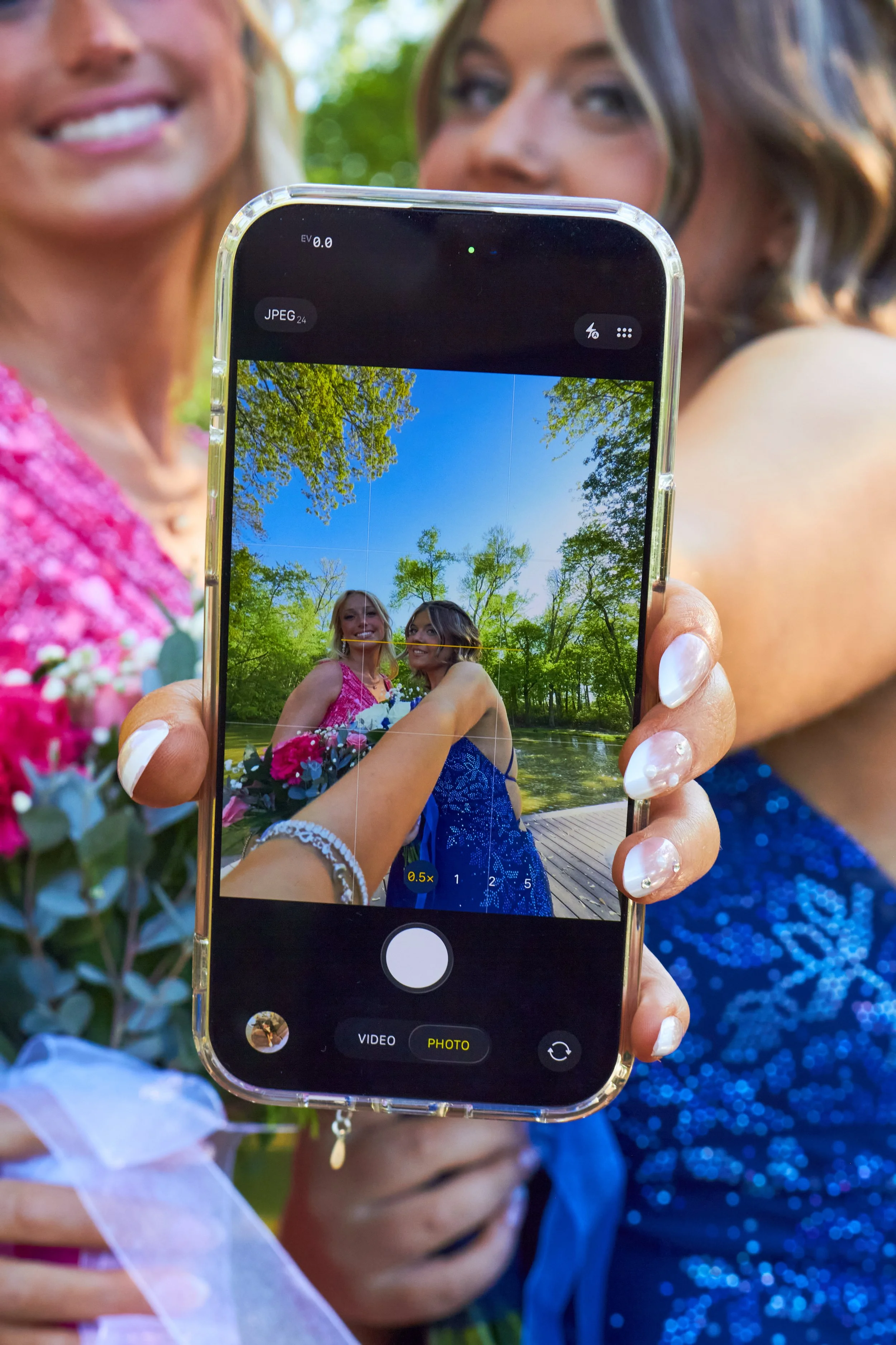 A person holding a smartphone taking a photo of two women outdoors near a lake, surrounded by green trees. One woman is wearing a pink dress and the other a blue dress.