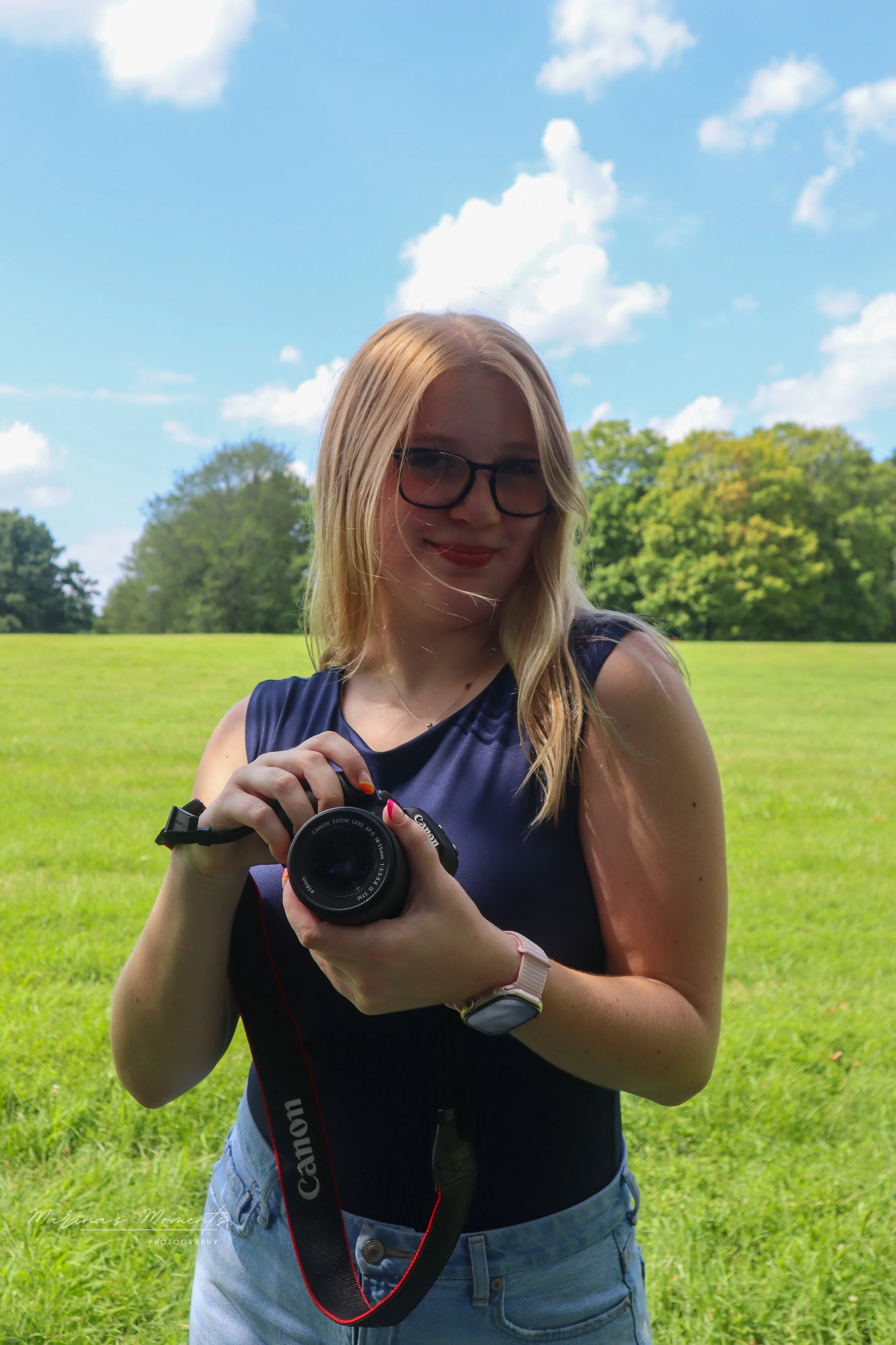 A young woman with blonde hair, glasses, and a navy blue sleeveless top holding a camera outdoors in a grassy field with trees and a blue sky with white clouds in the background.