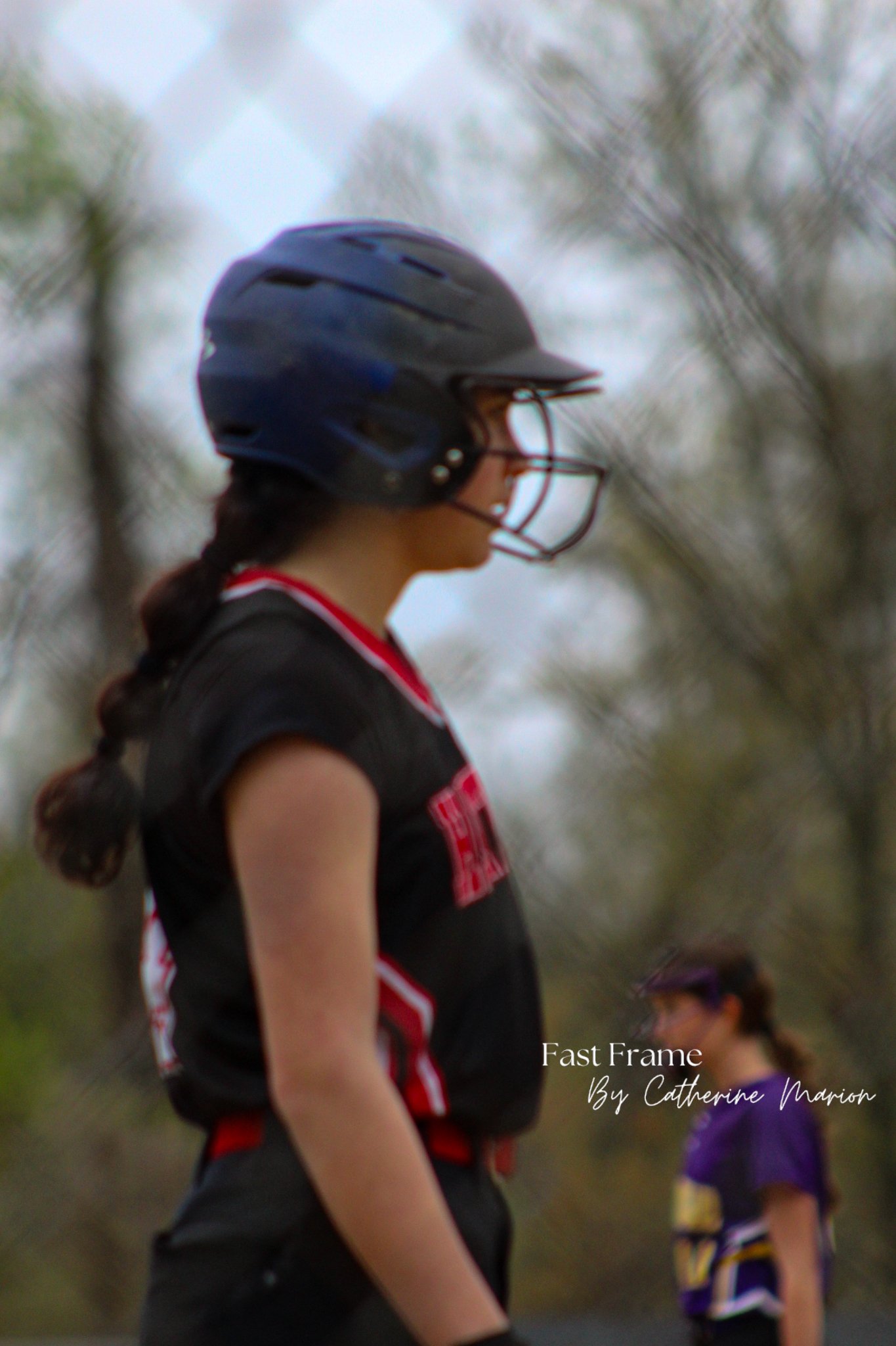 A girl with braided hair wearing a black sports shirt and a blue helmet, standing outdoors with trees in the background.