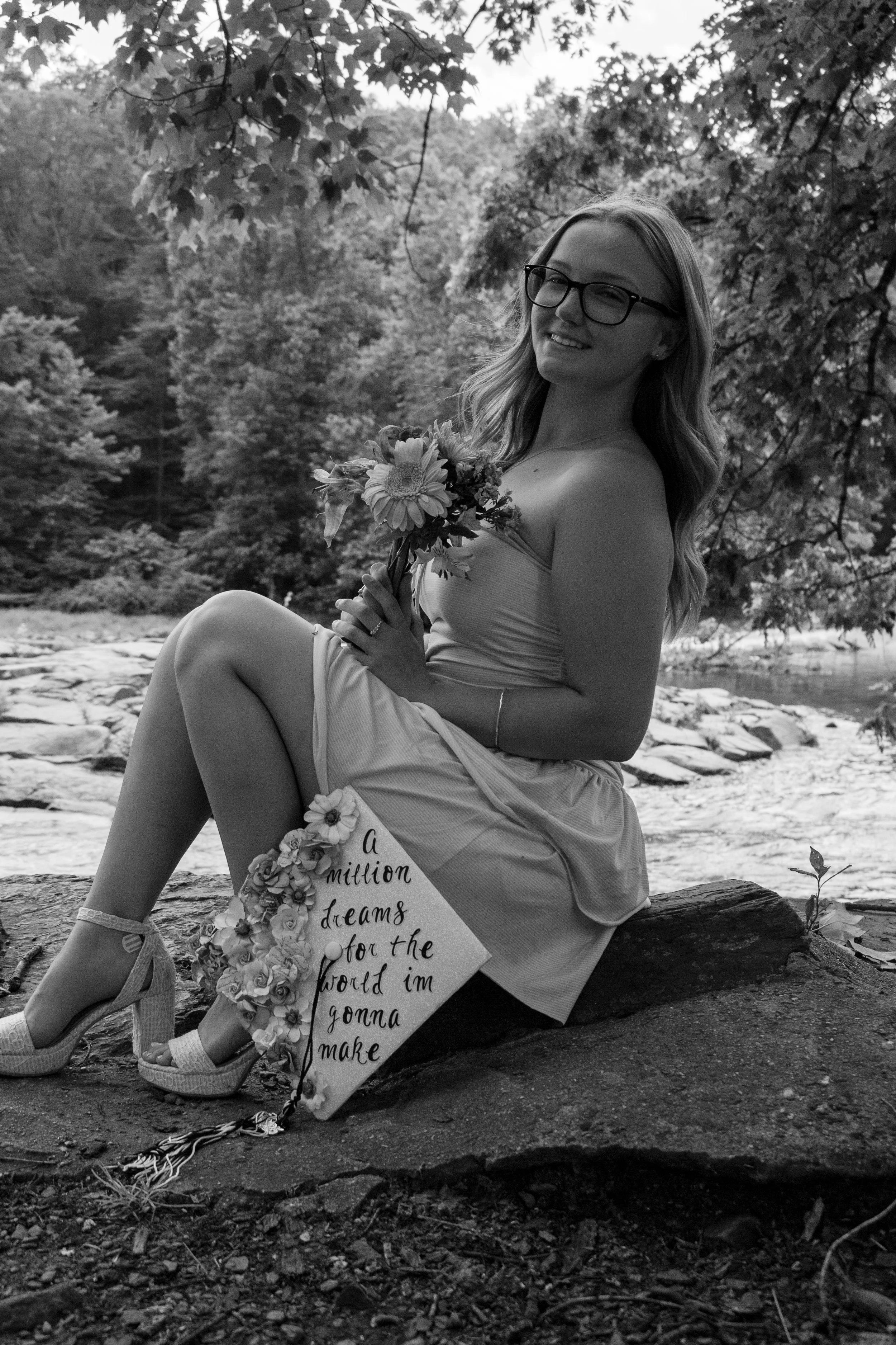 A young woman with glasses and long hair, sitting on a rock by a river in a wooded area, holding a bouquet of flowers and a small cake, smiling at the camera. There is a sign with a quote partially visible in front of her.