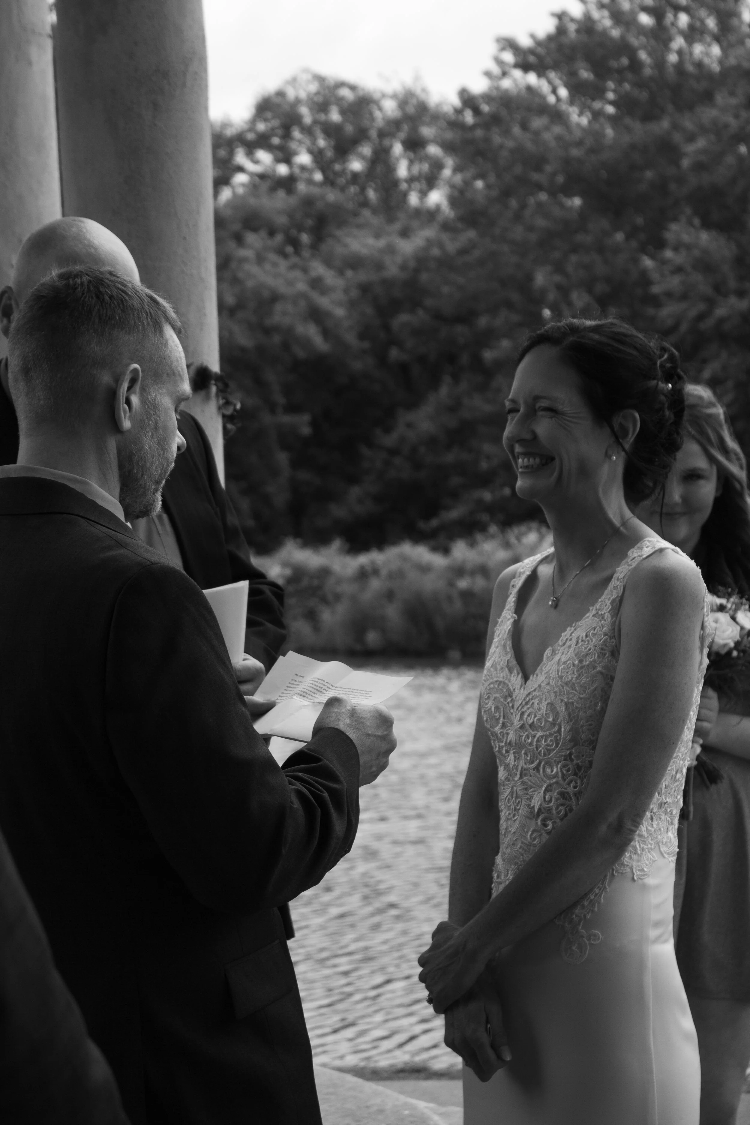 A black and white photo of a wedding ceremony outside by a body of water. The bride is smiling and wearing a lace wedding dress, and the groom is holding a paper, possibly reading vows.