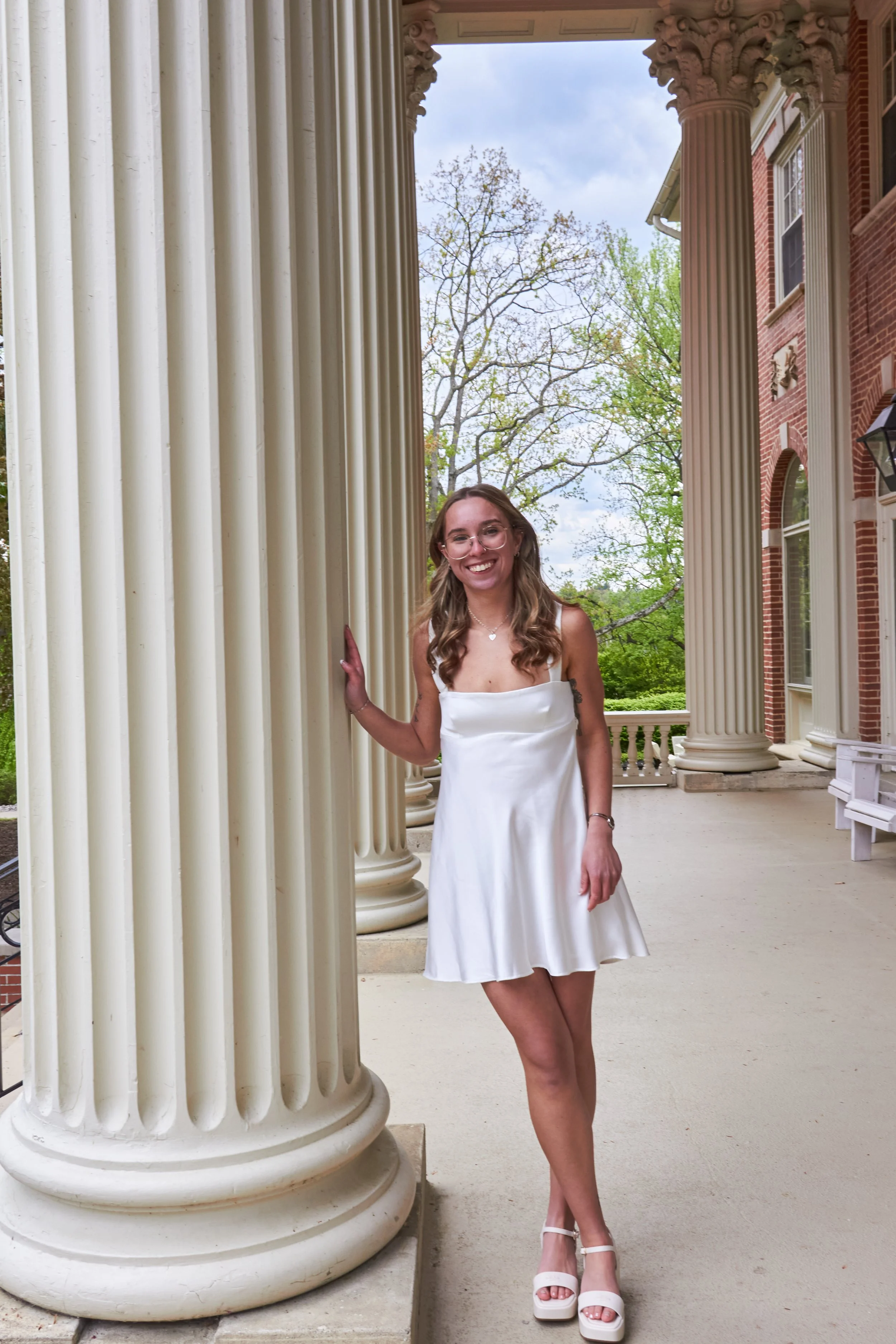 A young woman with long wavy hair, wearing glasses and a white satin dress with spaghetti straps, standing between large cream-colored classical columns on a porch of a red brick building. She is smiling and looking at the camera, with trees and a cl