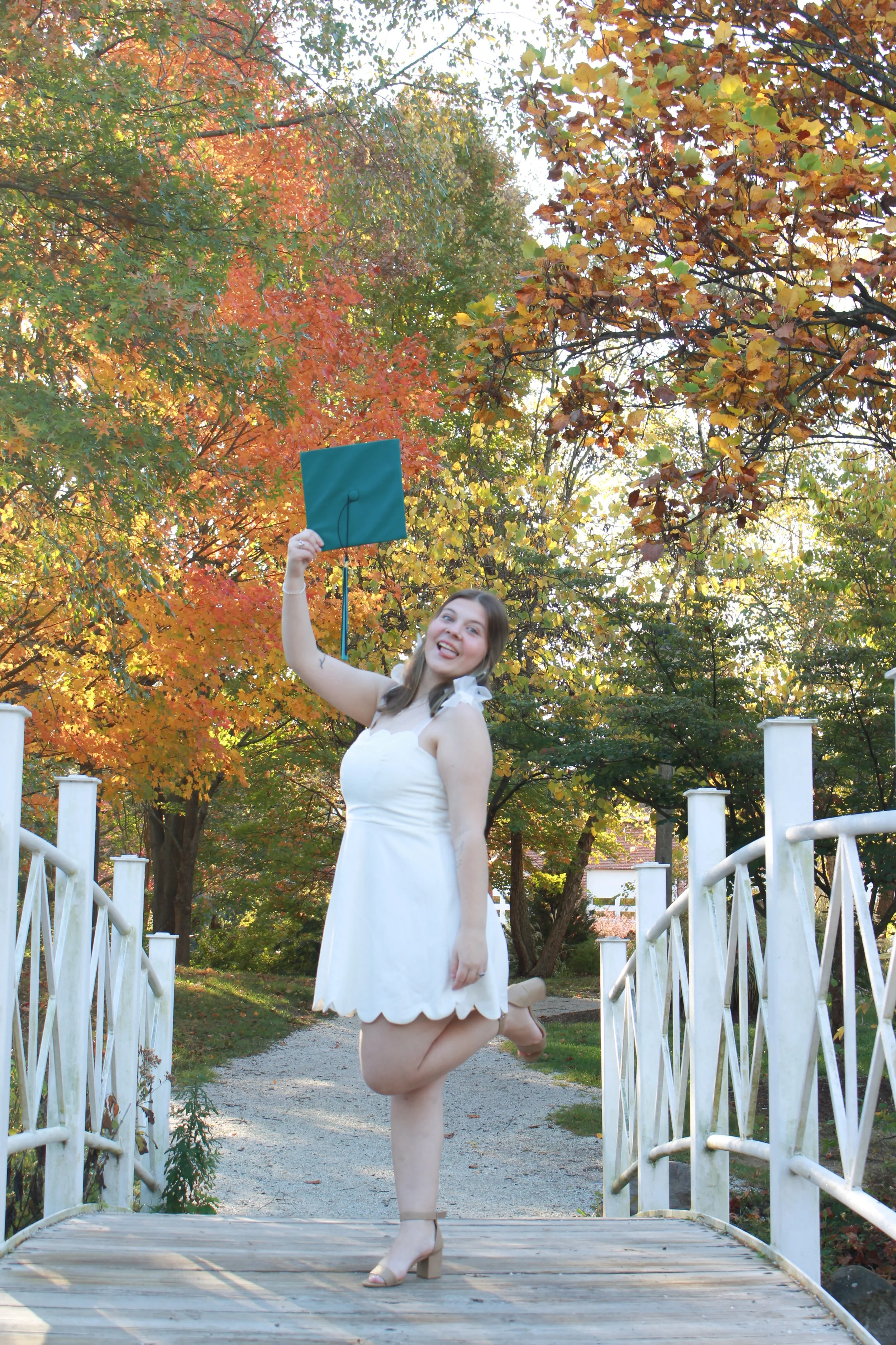 A woman in a white dress is kneeling on one leg on a small wooden bridge during autumn. She is smiling and holding a graduation cap in the air, with trees displaying fall foliage in the background.
