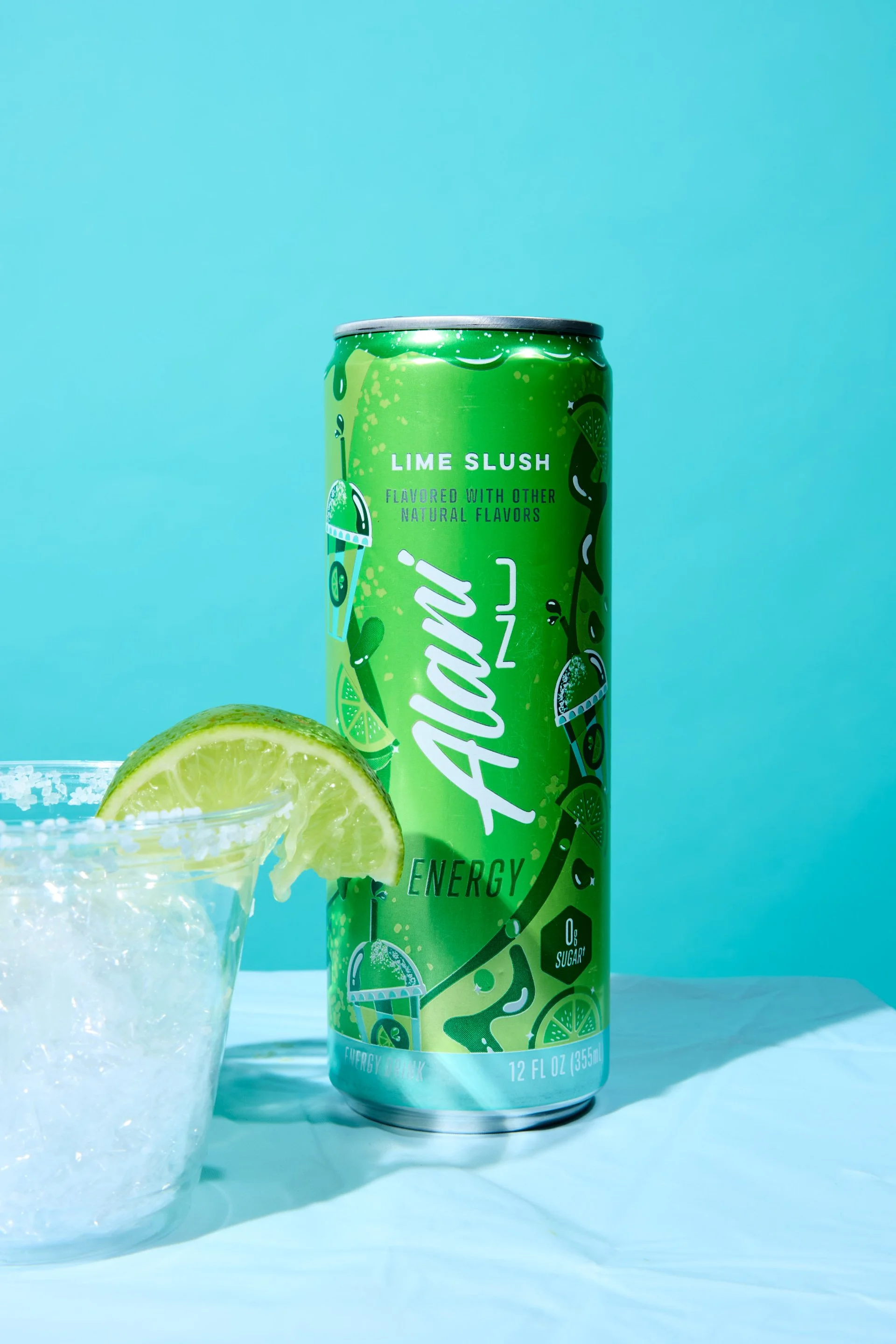 A green can of Alani Nu Lime Slush energy drink with a lemon wedge and a glass of water with lemon slices in the foreground on a light blue background.