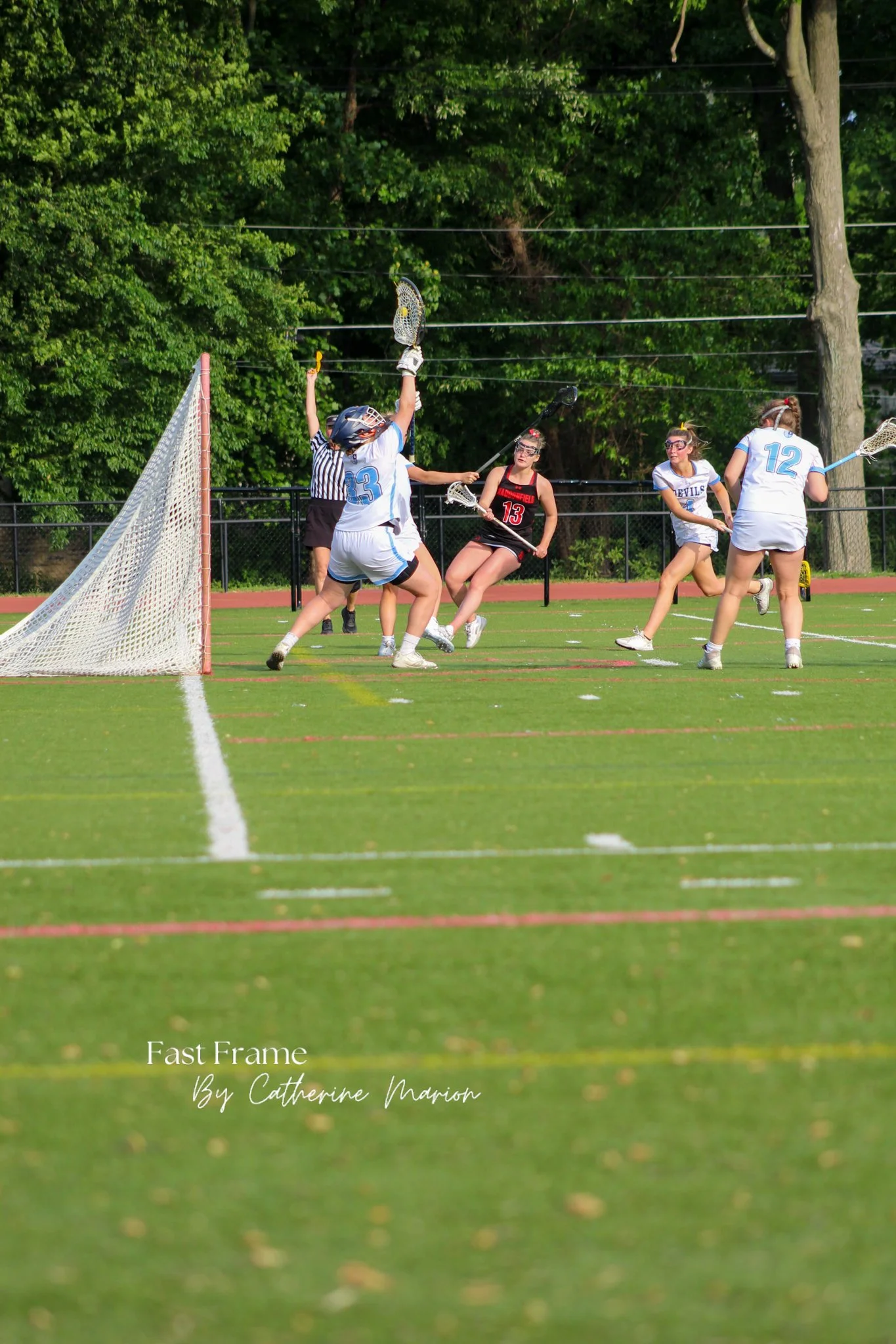 Young girls playing lacrosse on a grassy field, with some jumping and others holding lacrosse sticks, near a goal post, with trees and a fence in the background.
