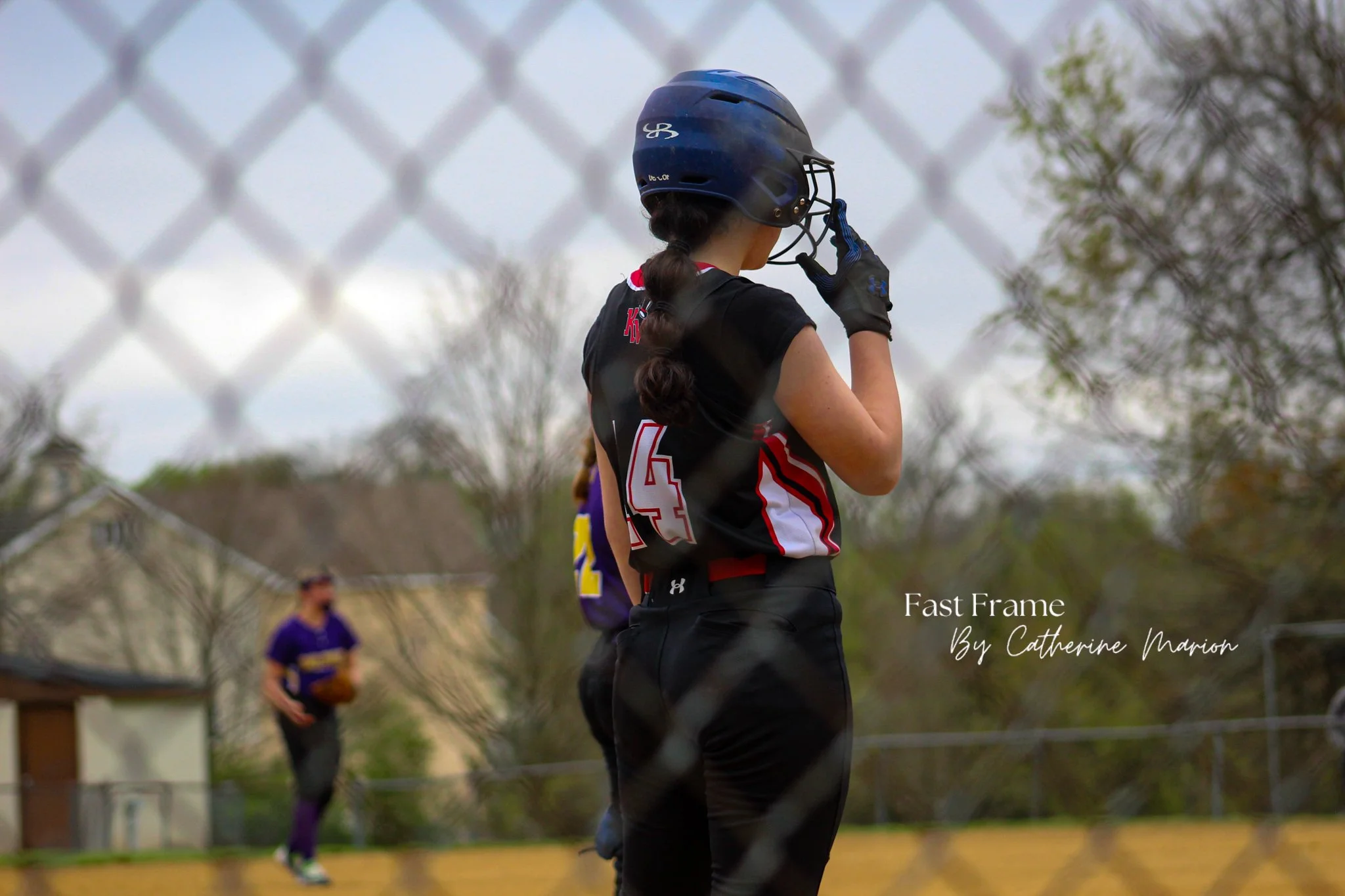A young female softball player wearing a dark uniform, helmet, and gloves stands on the field near a chain-link fence, with a blurred background of other players and trees.