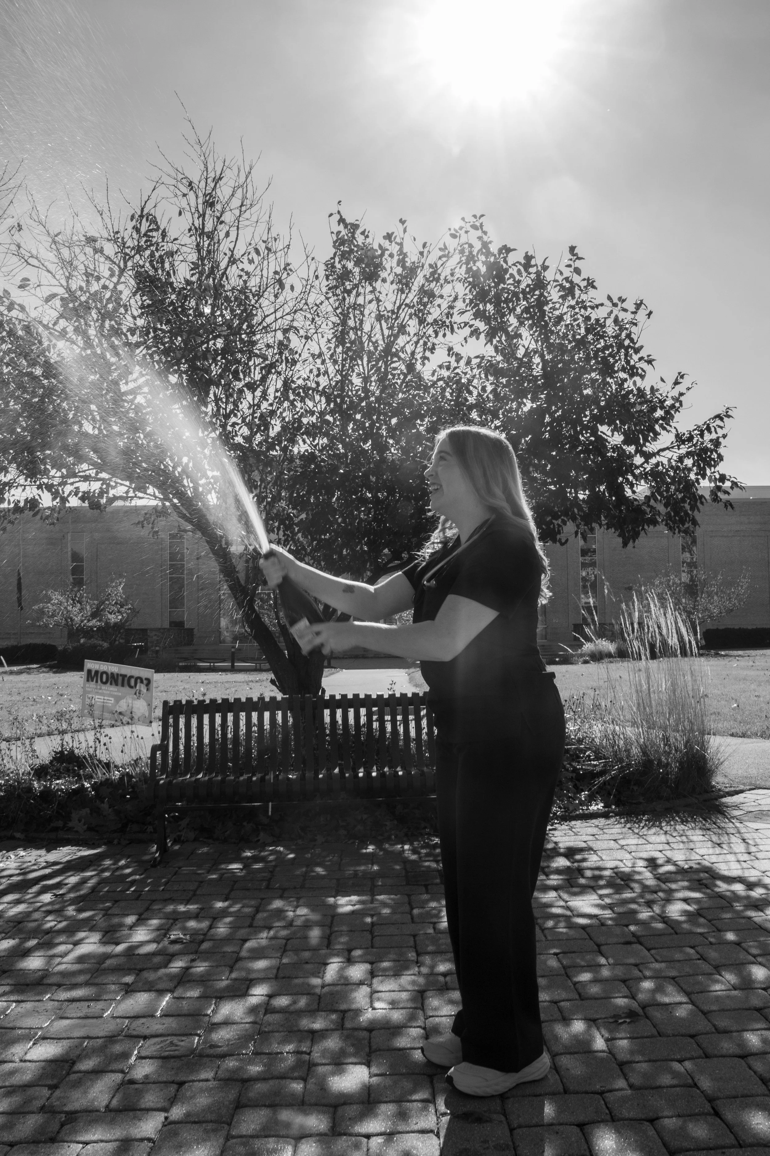 A woman standing outdoors in daylight, smiling and holding a rainbow-colored water spray, with a large tree and a building in the background. The ground is paved with bricks and there is a small fence and a sign nearby.