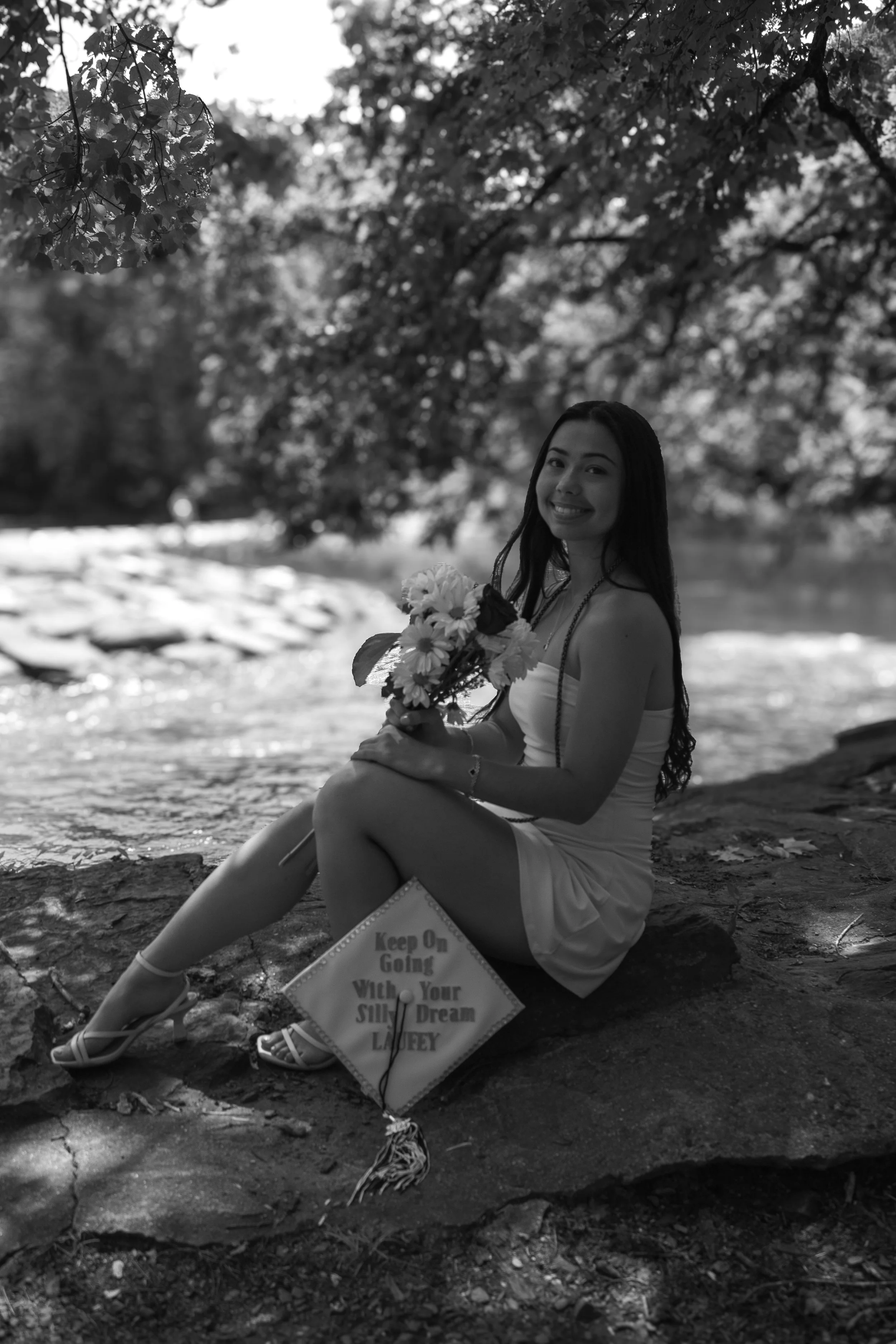 A smiling woman in a dress and heels sitting on a rock by a river, holding a bouquet of flowers, with trees overhead and a sign that reads 'Keep On Going With Your Silly Dream LADY'.