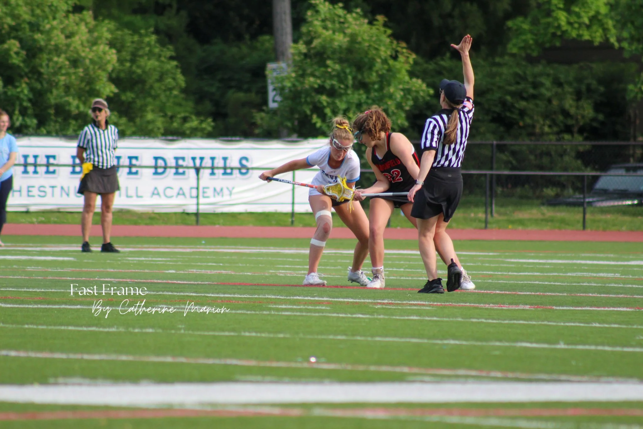 Two girls are competing in a lacrosse game, with one girl in a white jersey and the other in a black jersey, on a green field with a race track and trees in the background. A female referee in a black and white striped shirt is signaling with her han