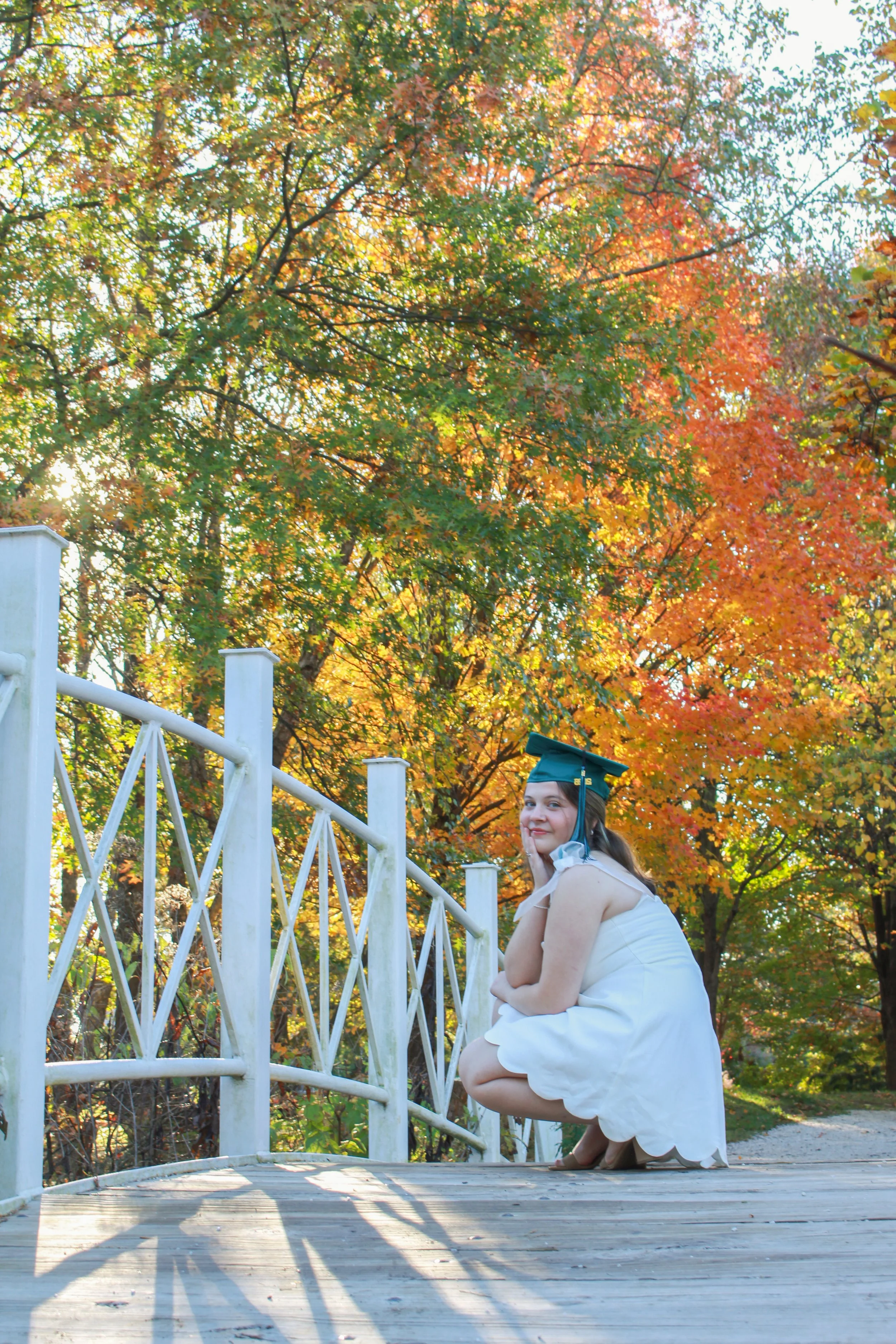 A young woman in a white dress and green graduation cap crouches on a wooden bridge with metal railing, surrounded by colorful autumn trees with orange, yellow, and green leaves.