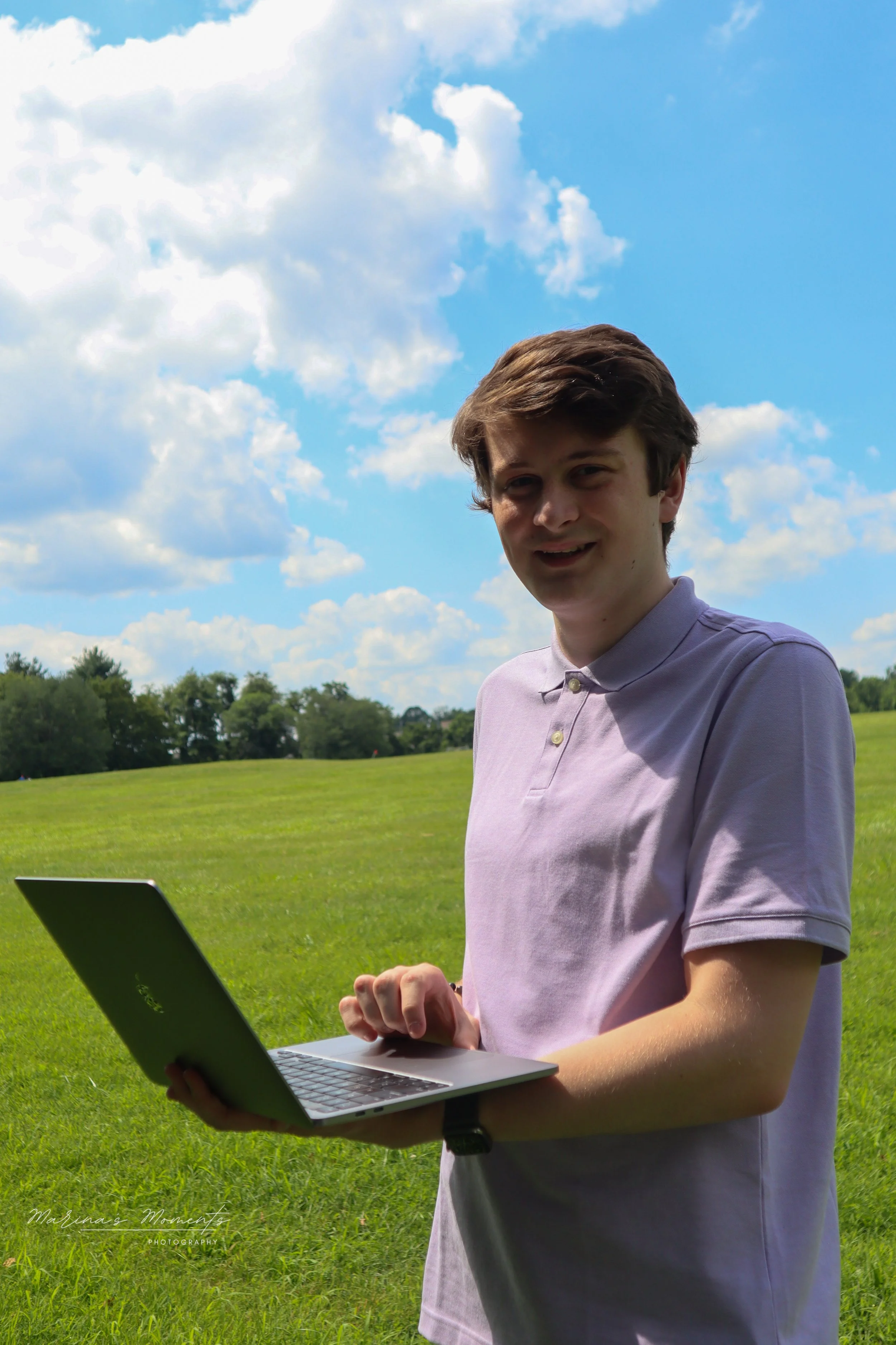 Young man with brown hair in a lavender polo shirt using a laptop outdoors on a grassy field with trees and a partly cloudy sky in the background.